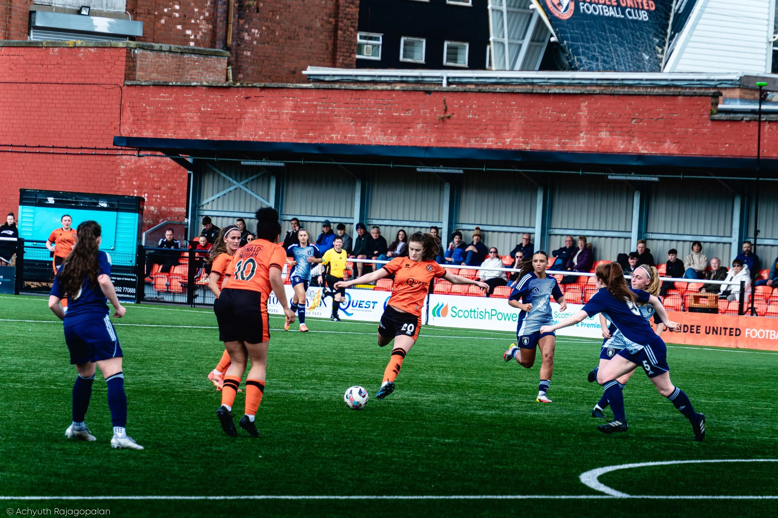 Women playing soccer on a green field with spectators watching from behind a fence and a building with a sign reading 'Coventry United Football Club' in the background.