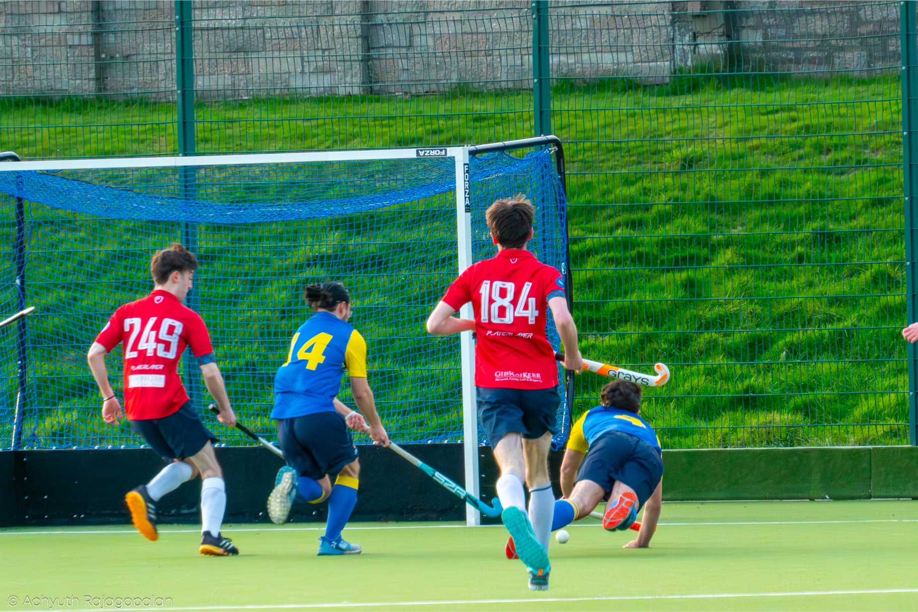 Children playing field hockey during a game, with one player in a yellow and blue uniform falling, and other players around the goal, with a metal fence and green grassy background.