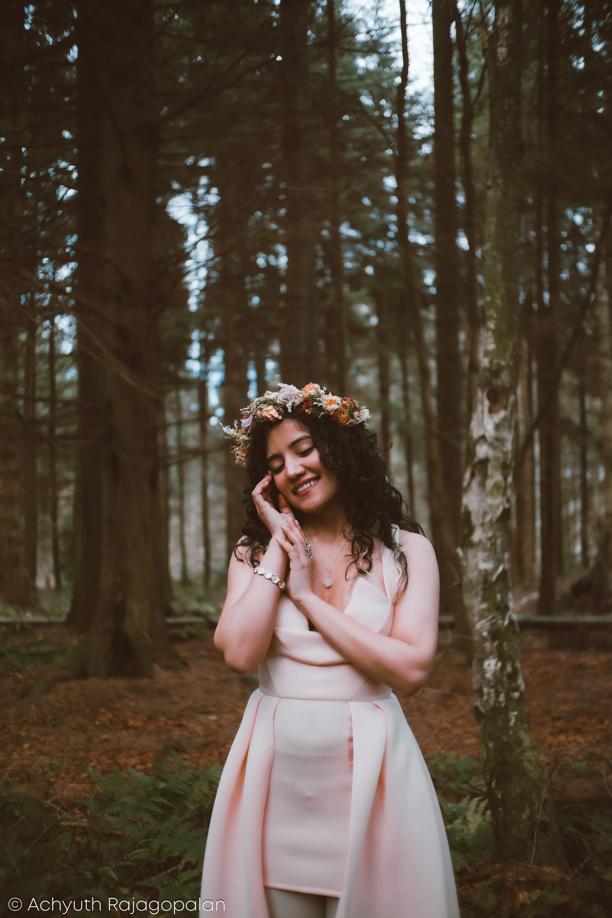 A woman with curly dark hair wearing a floral crown and a satin dress, standing in a forested area with tall trees and green foliage, smiling and looking down with her hand near her face.