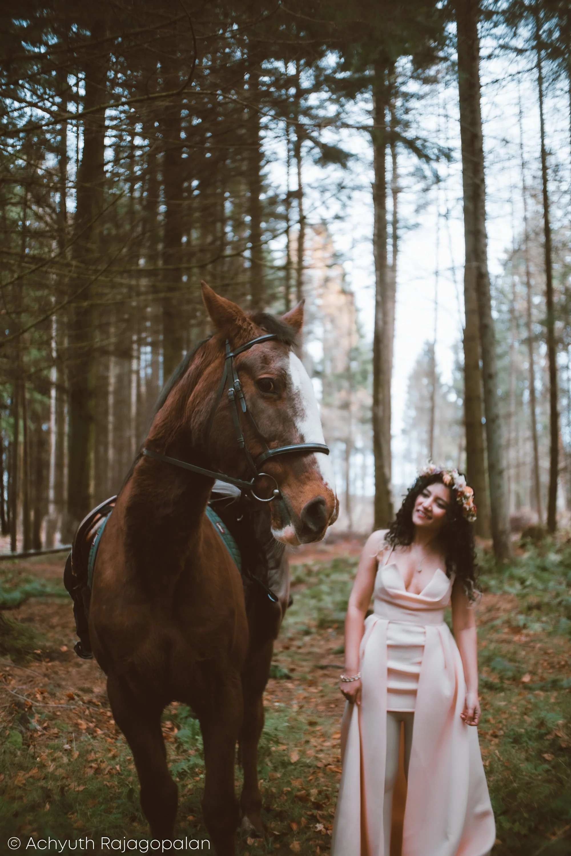 A woman wearing a white dress and a flower crown stands in a forest, smiling at a brown horse in front of her. The horse is equipped with a bridle and saddle, and the woman appears joyful.