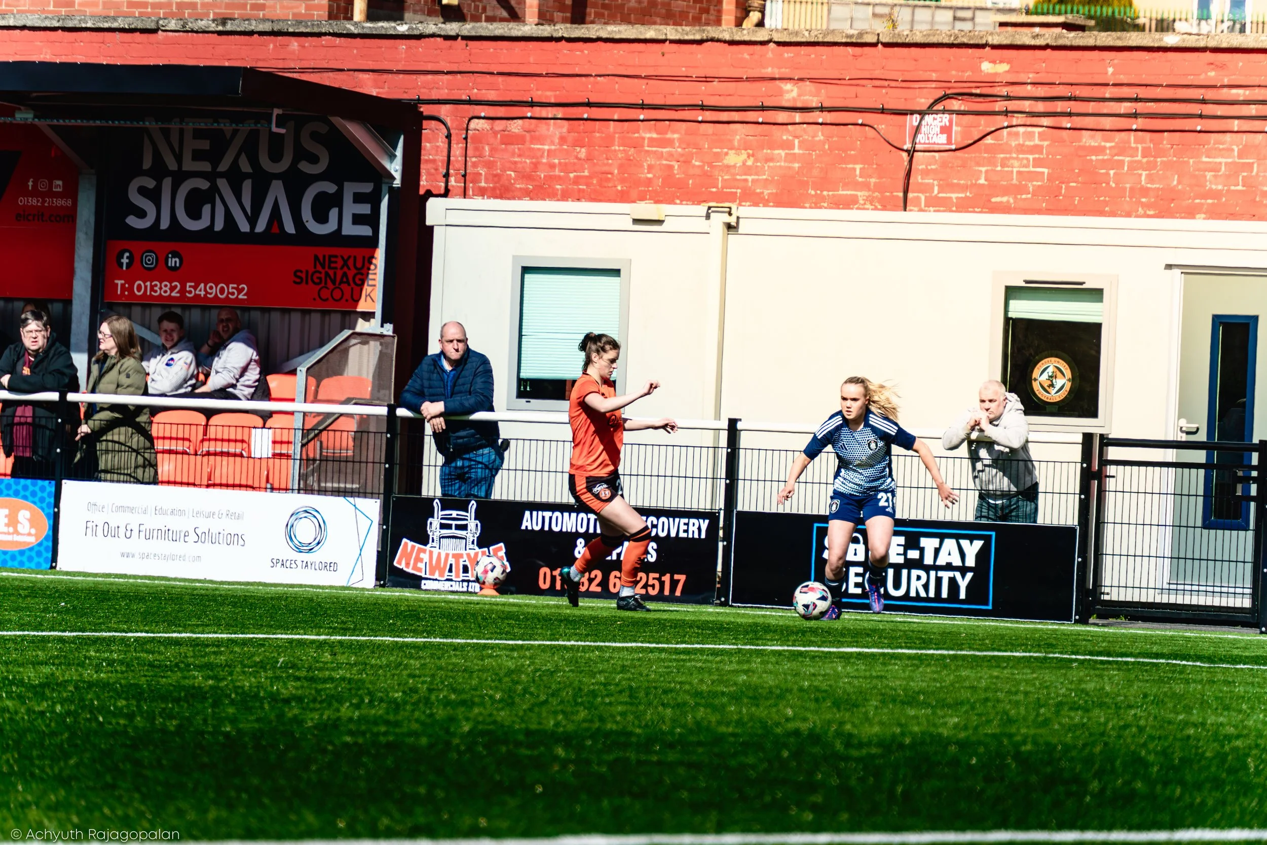 People watching a soccer game from the sidelines of a field with advertisements and a red brick building in the background.