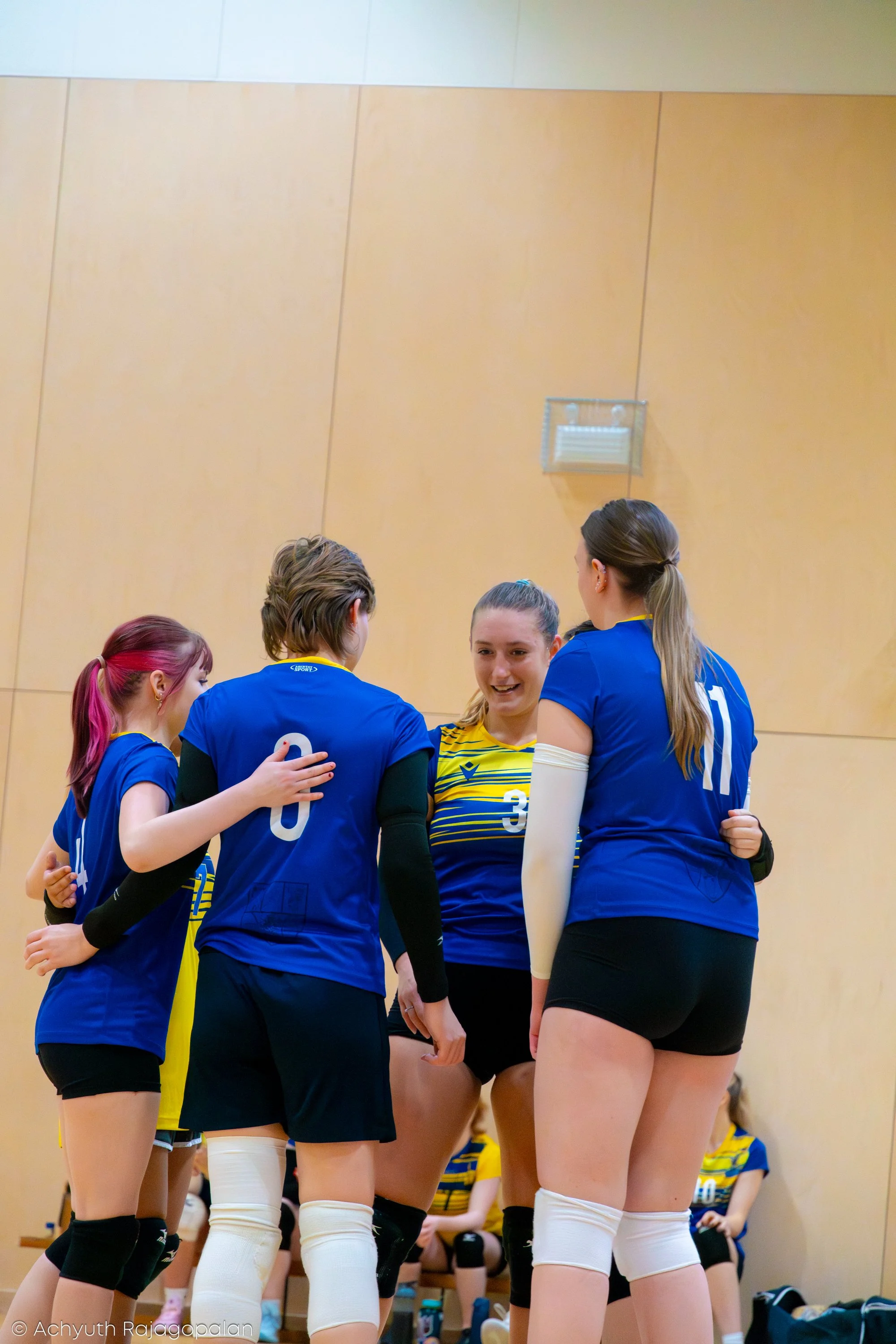 A group of female volleyball players in blue uniforms huddles together in a gymnasium, likely during a game or practice.