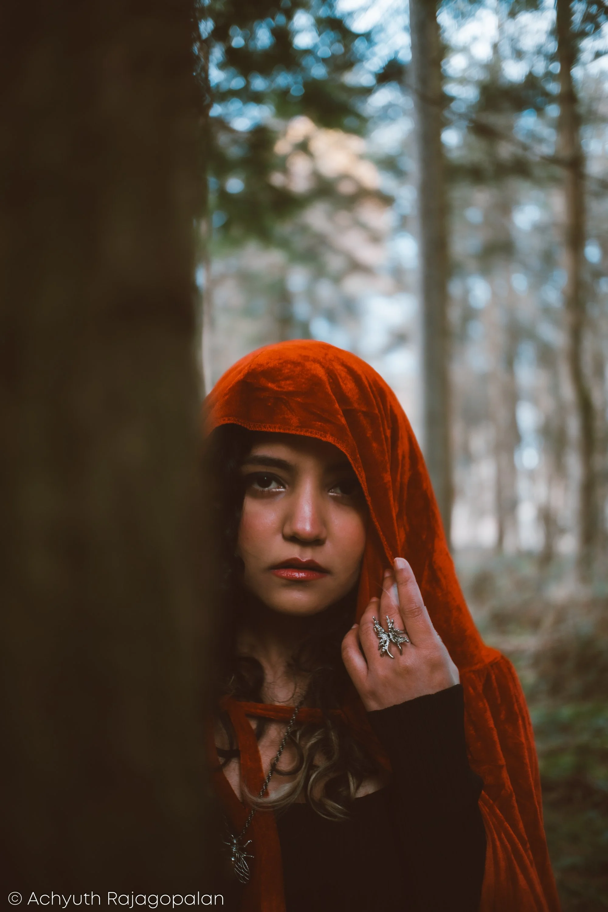 Woman with dark hair and brown eyes wearing a red hooded cloak, standing outdoors in a forest, touching her face with her left hand, which has a silver dragon ring. Trees and a cloudy sky are in the background.