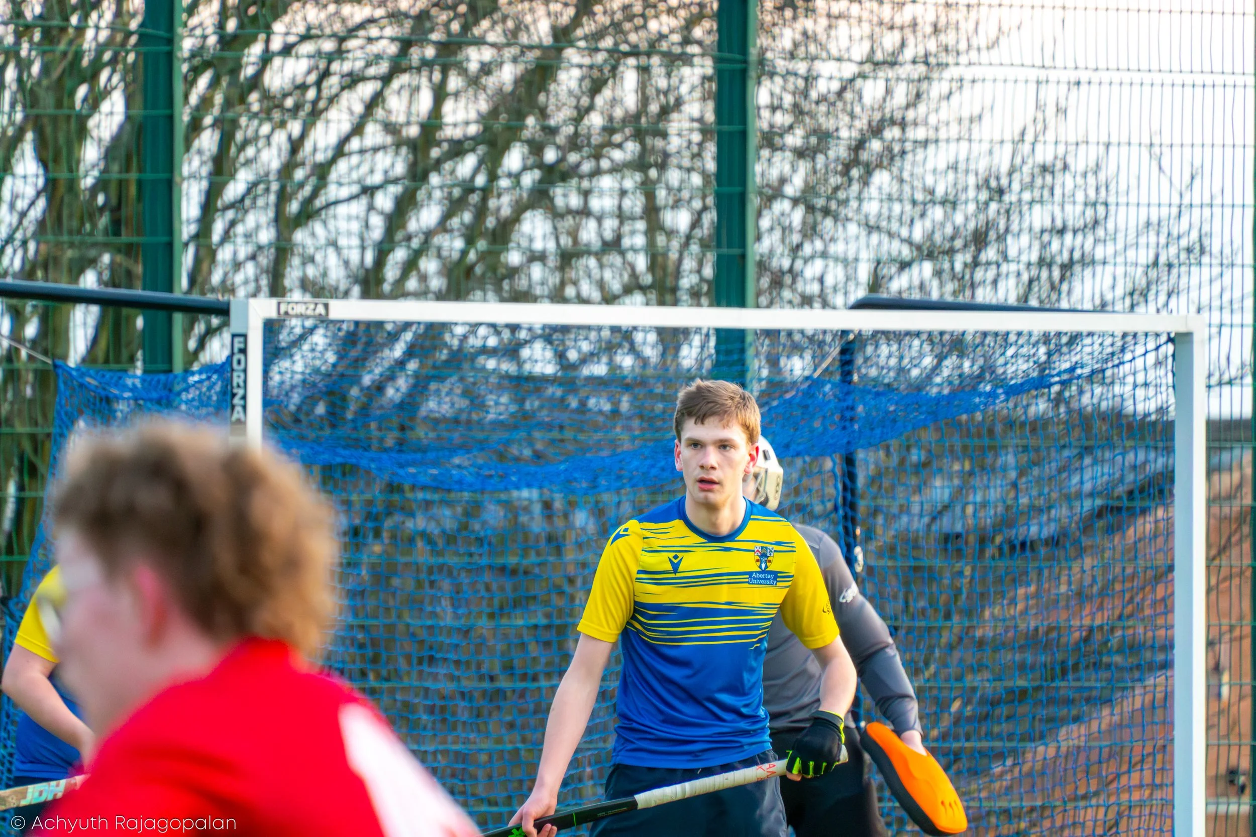 Hockey goalkeeper in a yellow and blue uniform standing in front of a goal net during a match, with players and a fence with trees in the background.