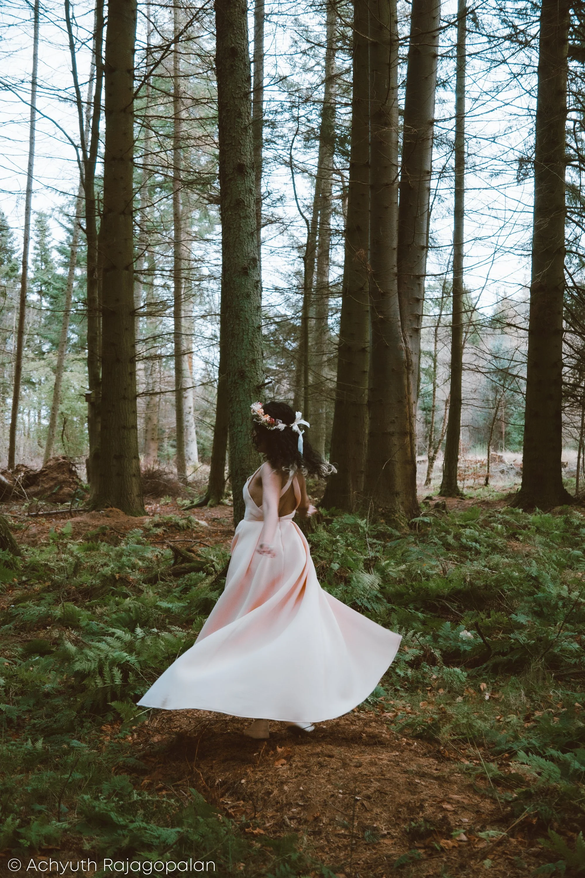 A woman in a flowing white gown with a floral crown is walking through a dense forest with tall, thin trees and green ferns on the ground.