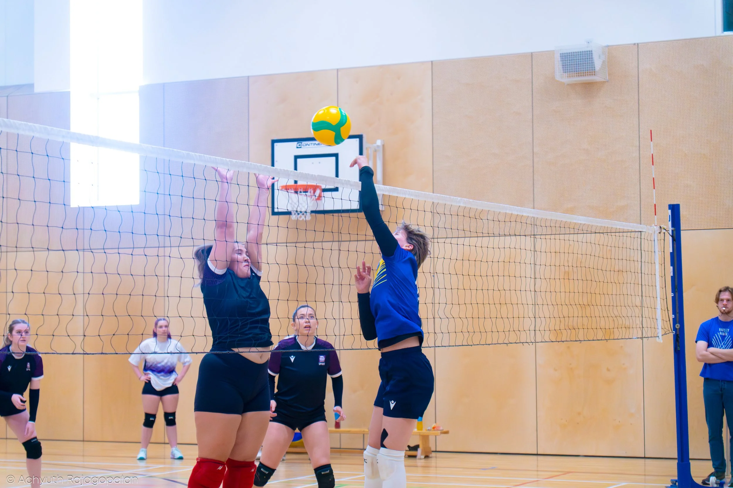 Two volleyball players jump to hit the ball at the net during a game in an indoor gymnasium, with several spectators watching.