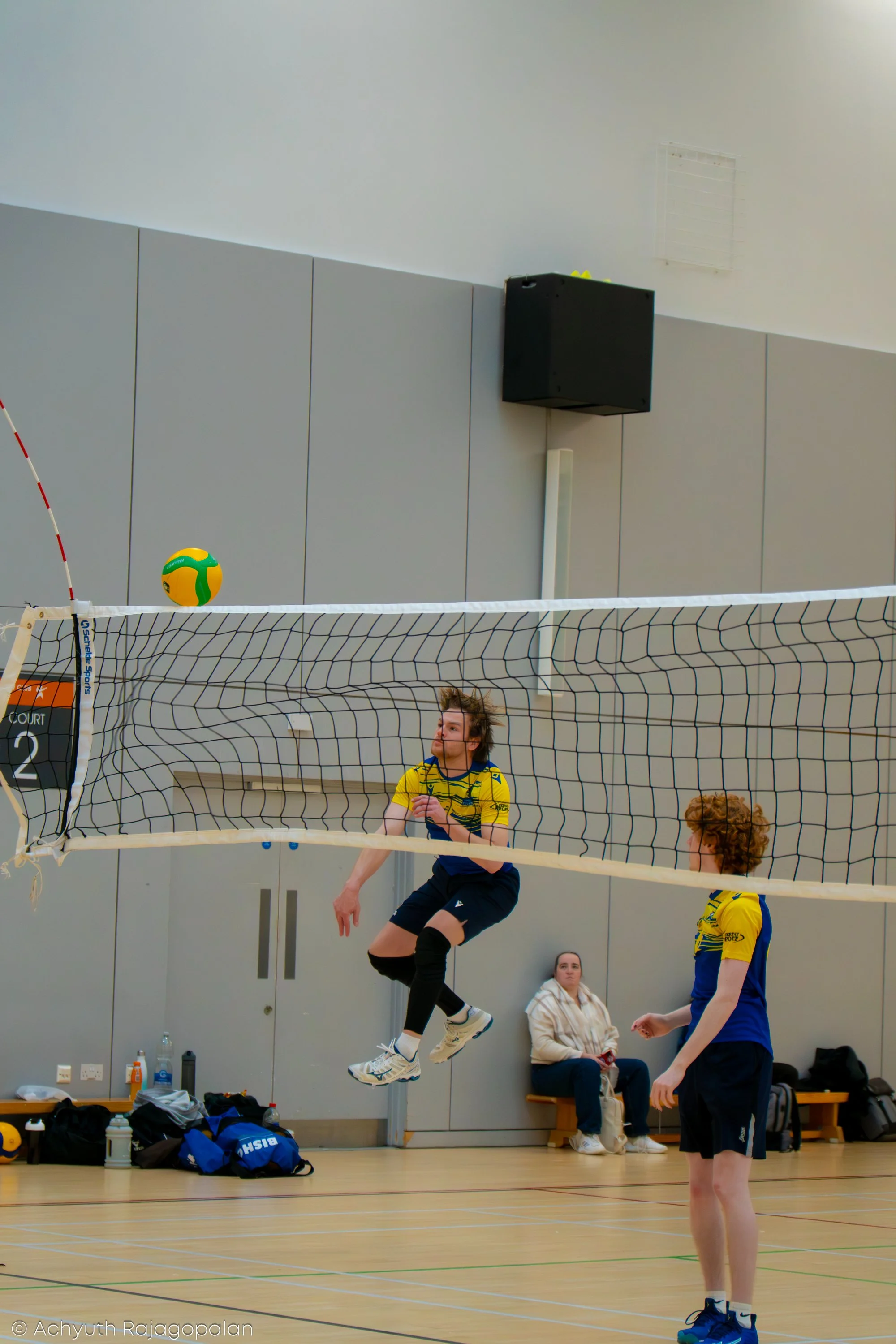 A volleyball game in progress with players jumping near the net and a ball in mid-air. Spectators are seated along the wall watching the game.