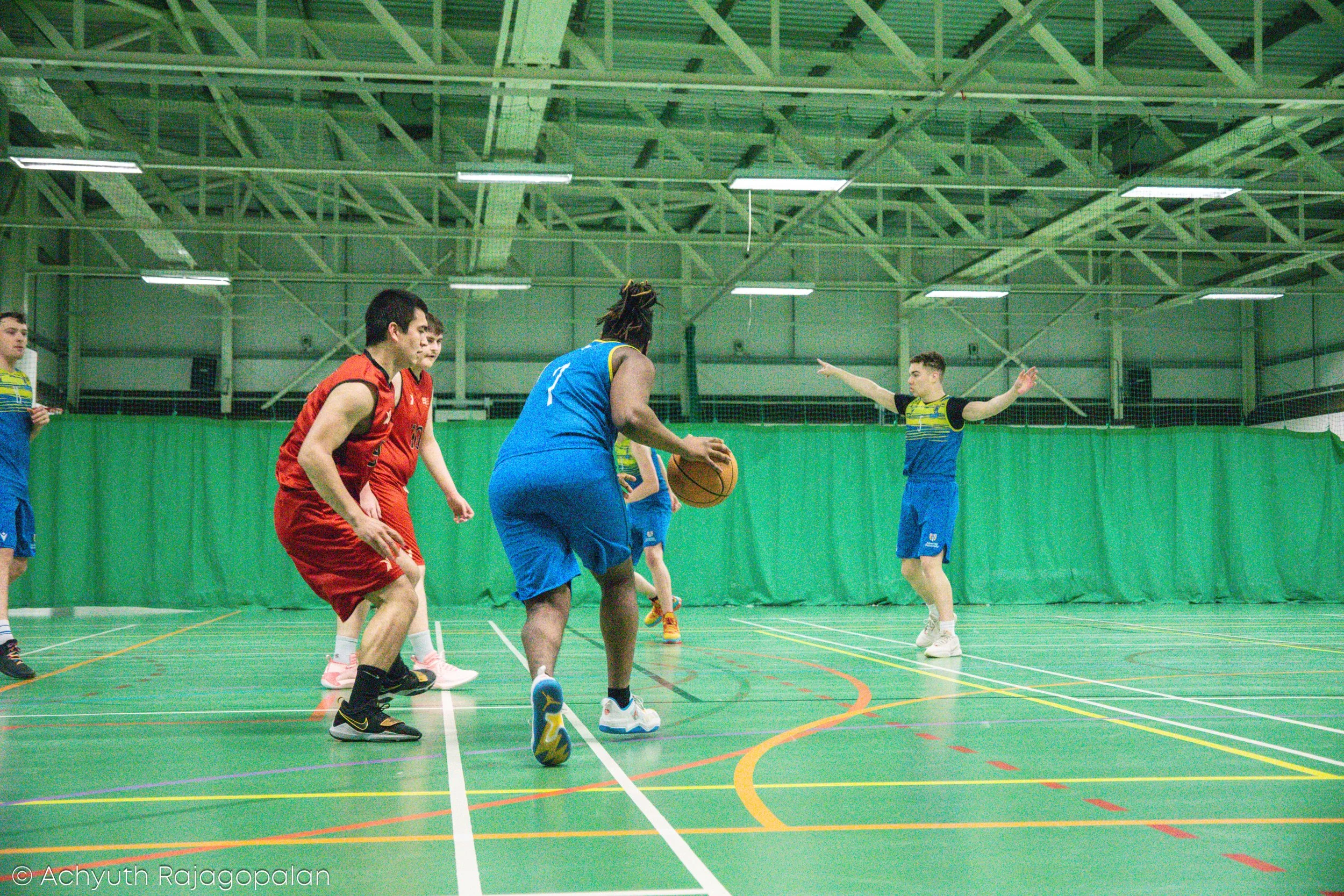 A group of young men playing basketball indoors on a green court with a green curtain in the background. One player in a blue jersey is holding the basketball, surrounded by players in red and blue jerseys.