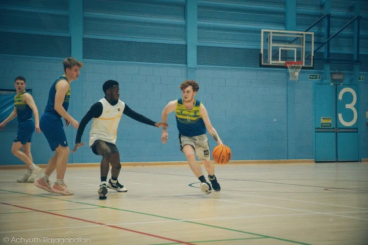 Young boys playing basketball in an indoor gym, with one boy dribbling the ball while others attempt to defend.