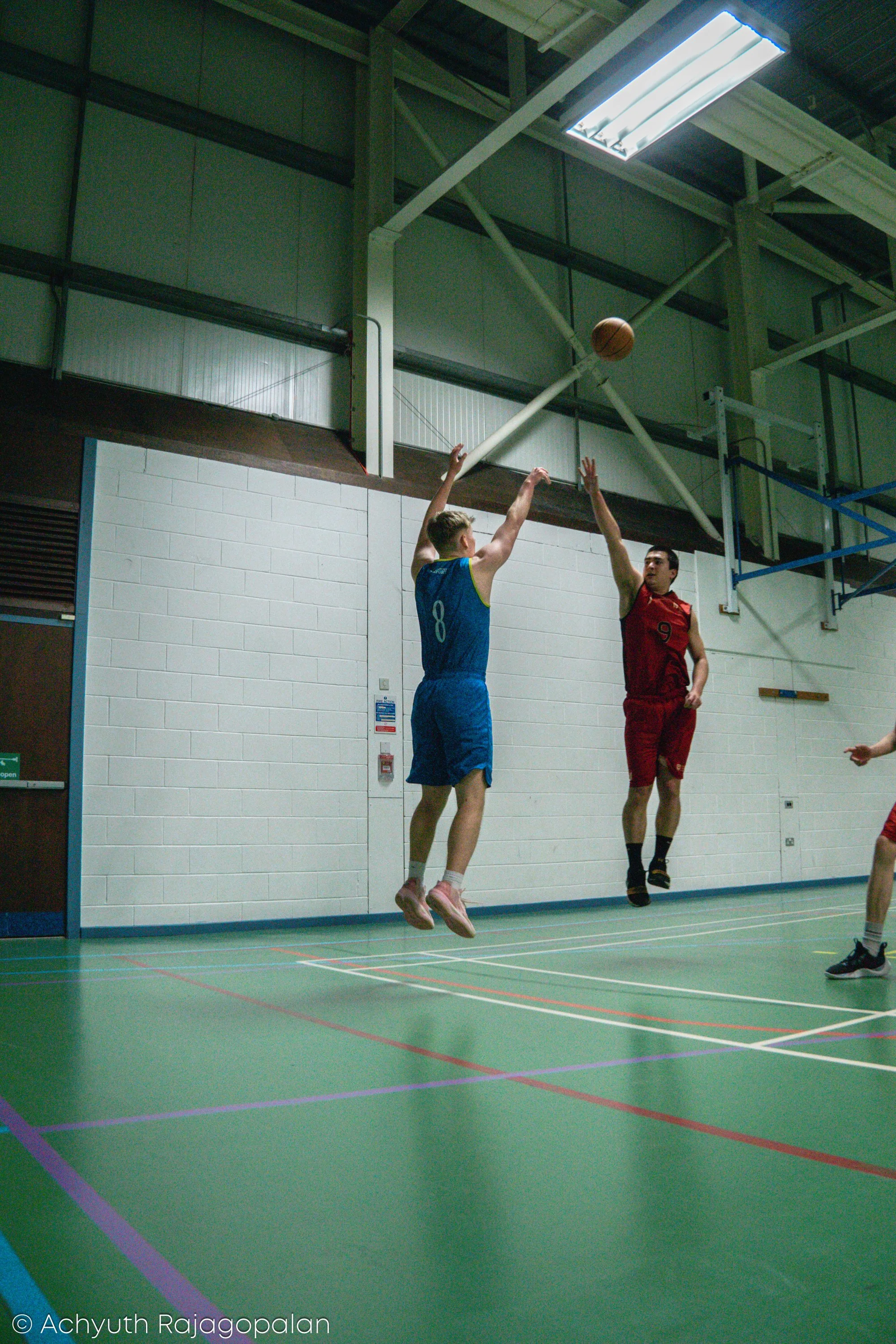 Two male volleyball players are jumping in the air to hit a volleyball during a game in an indoor gymnasium with green flooring and white brick walls.