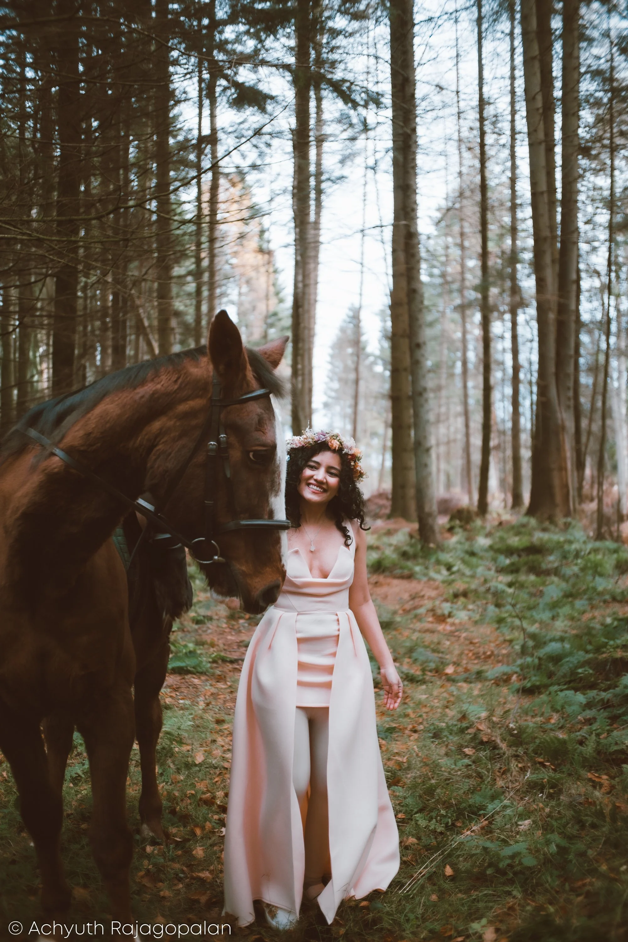 A smiling woman wearing a light-colored dress and floral crown, standing in a forest next to a brown horse with a black mane.