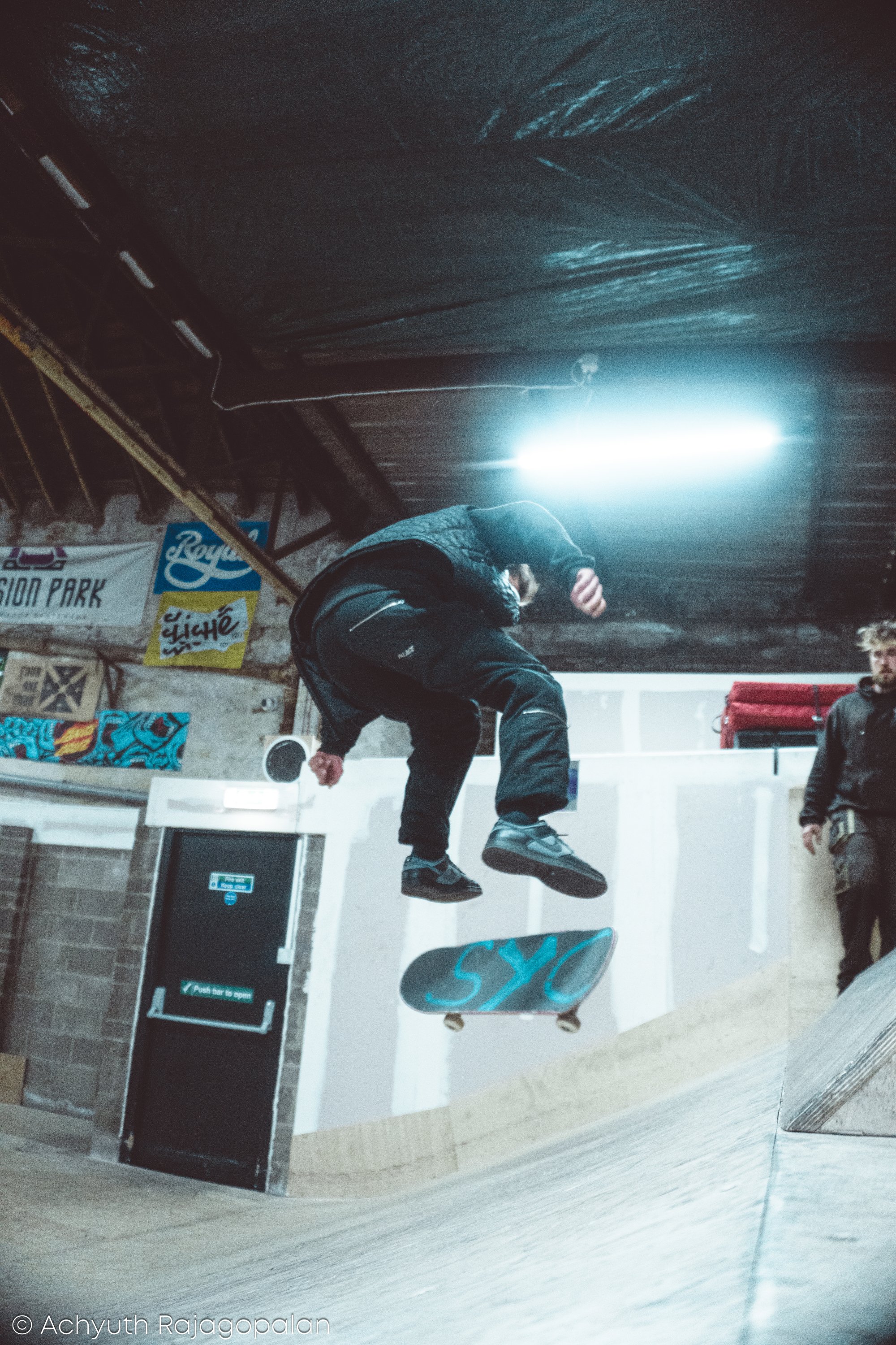 Person skateboarding indoors near a wall with posters, wearing a black jacket and pants, and with a skateboard with blue graphics.