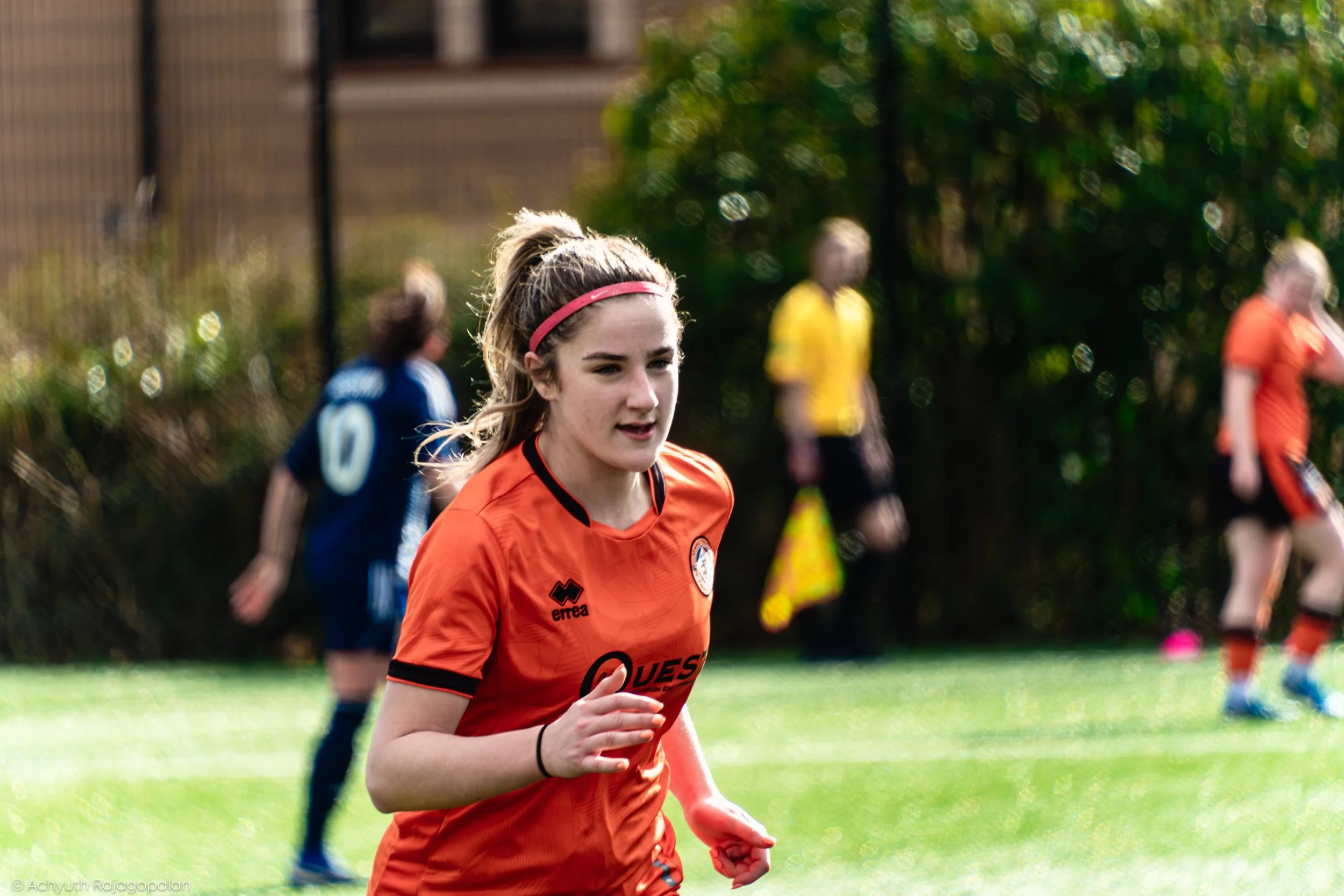 A young female soccer player with long blonde hair, wearing an orange jersey with black trims and a pink headband, jogging on a grassy field. Behind her, other players in yellow and blue jerseys are visible, slightly blurred, and there are trees and 