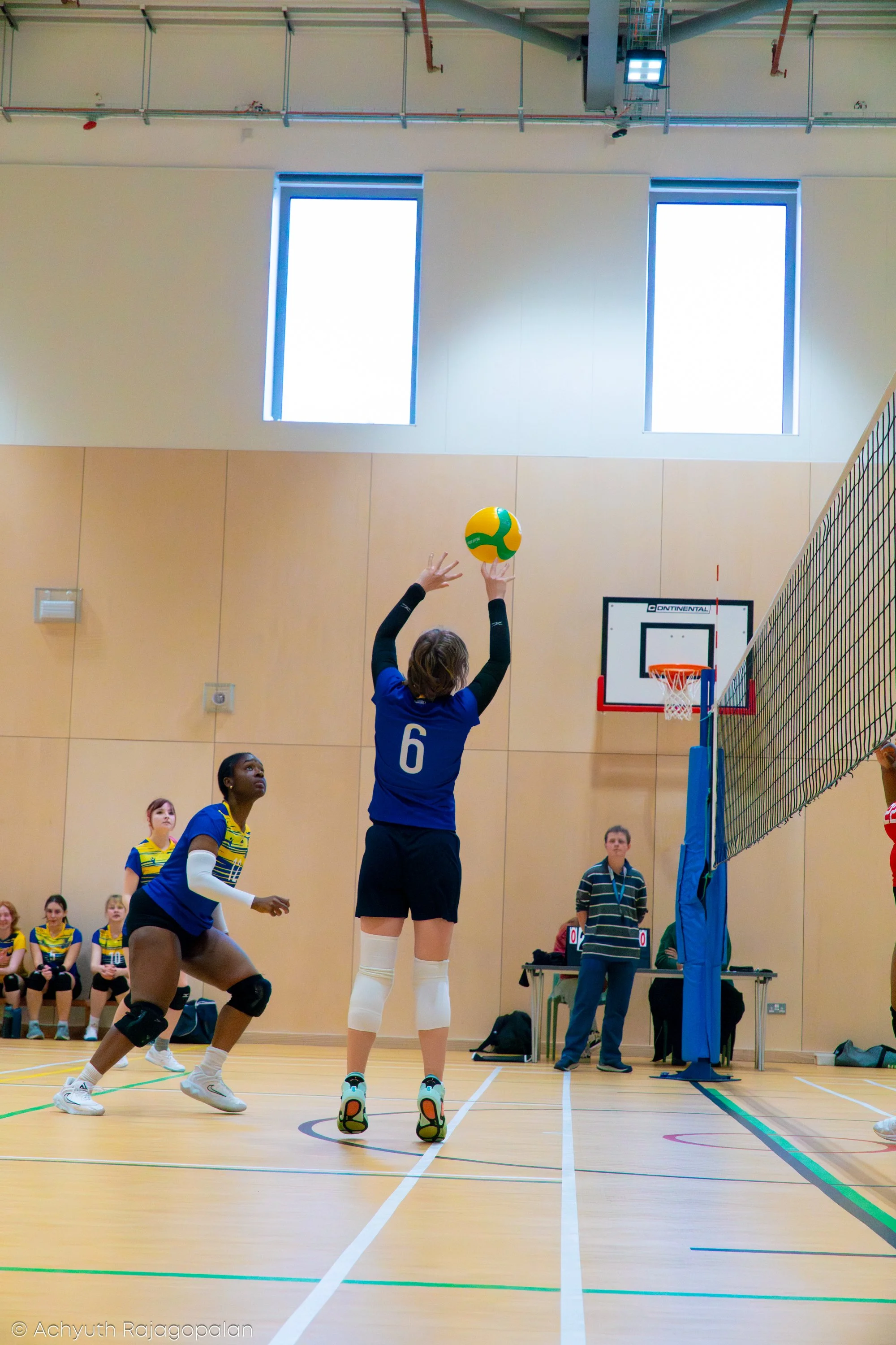 A volleyball game with players in a gymnasium. A player wearing a blue jersey with the number 6 is jumping to hit the volleyball over the net. Other players and spectators are visible in the background.