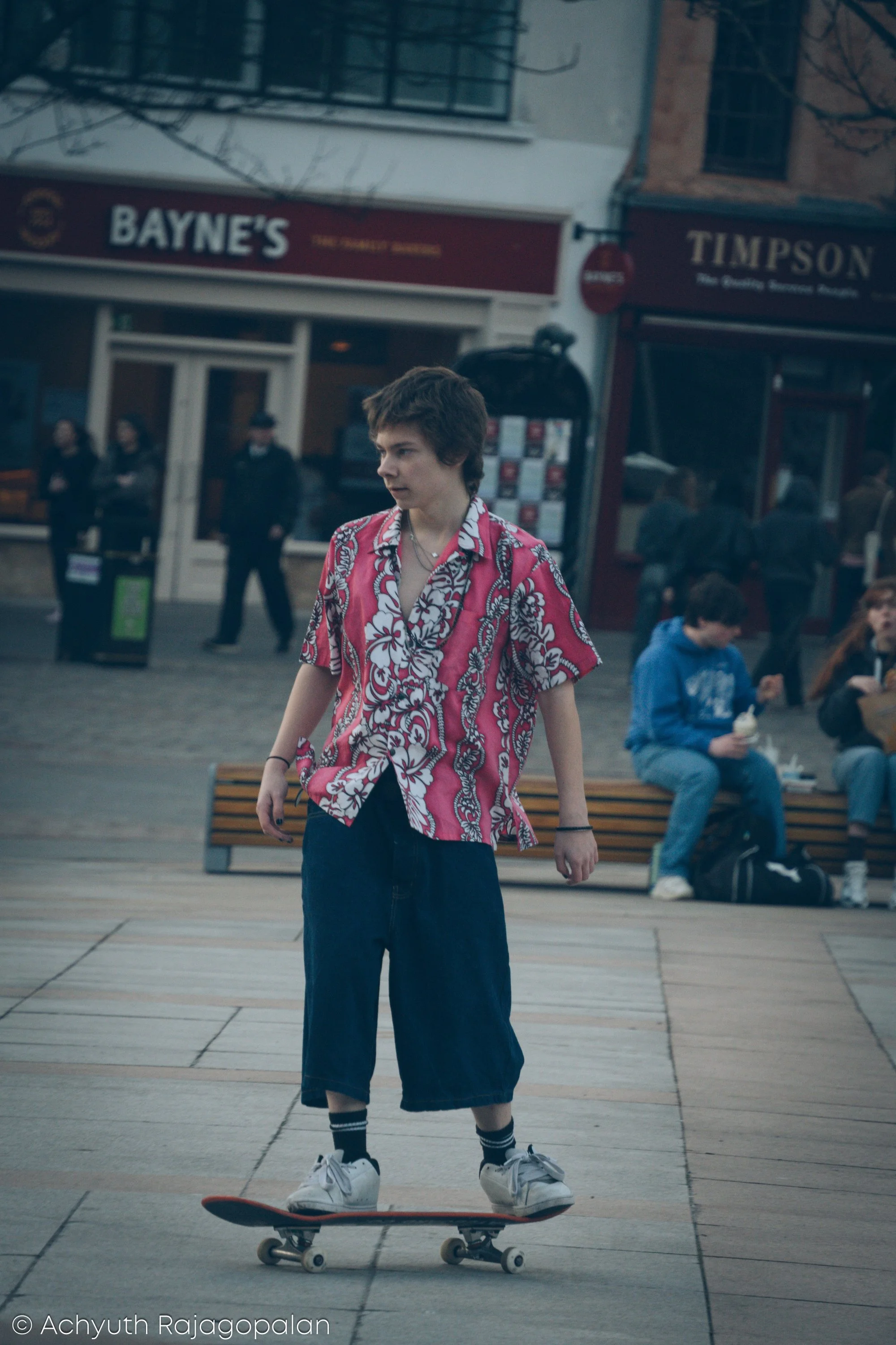 A young person skateboarding in an urban outdoor setting, wearing a bright pink Hawaiian shirt, dark wide-leg jeans, and white sneakers. There are people sitting on benches and standing in the background, with storefronts and buildings visible.