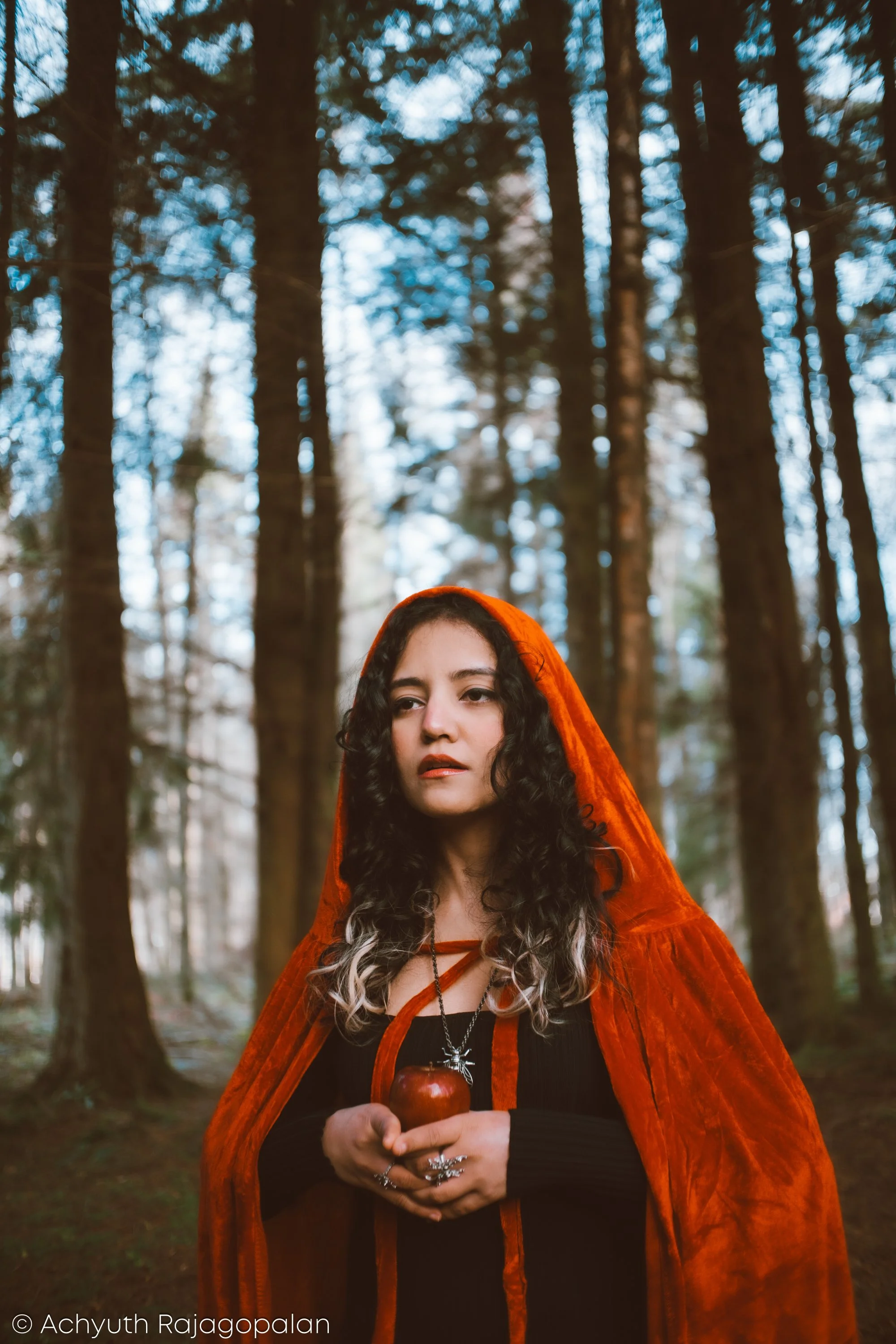 A woman with curly dark hair wearing an orange cloak and black clothing, holding a red apple and a necklace with a cross, standing in a forest with tall trees and sunlight filtering through.