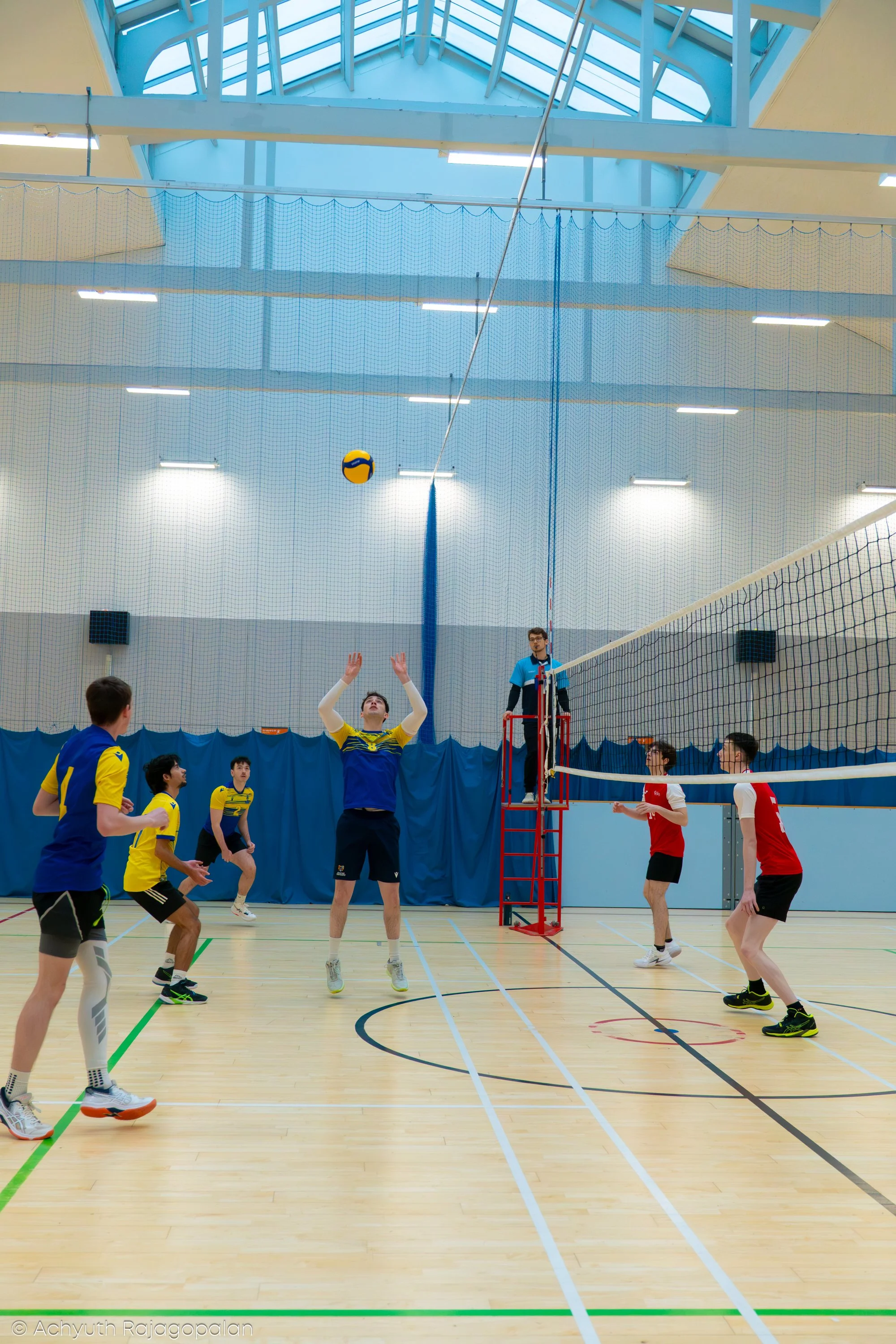 Indoor volleyball game in progress with players jumping to attempt to hit the ball over the net, with referees and spectators in the background.