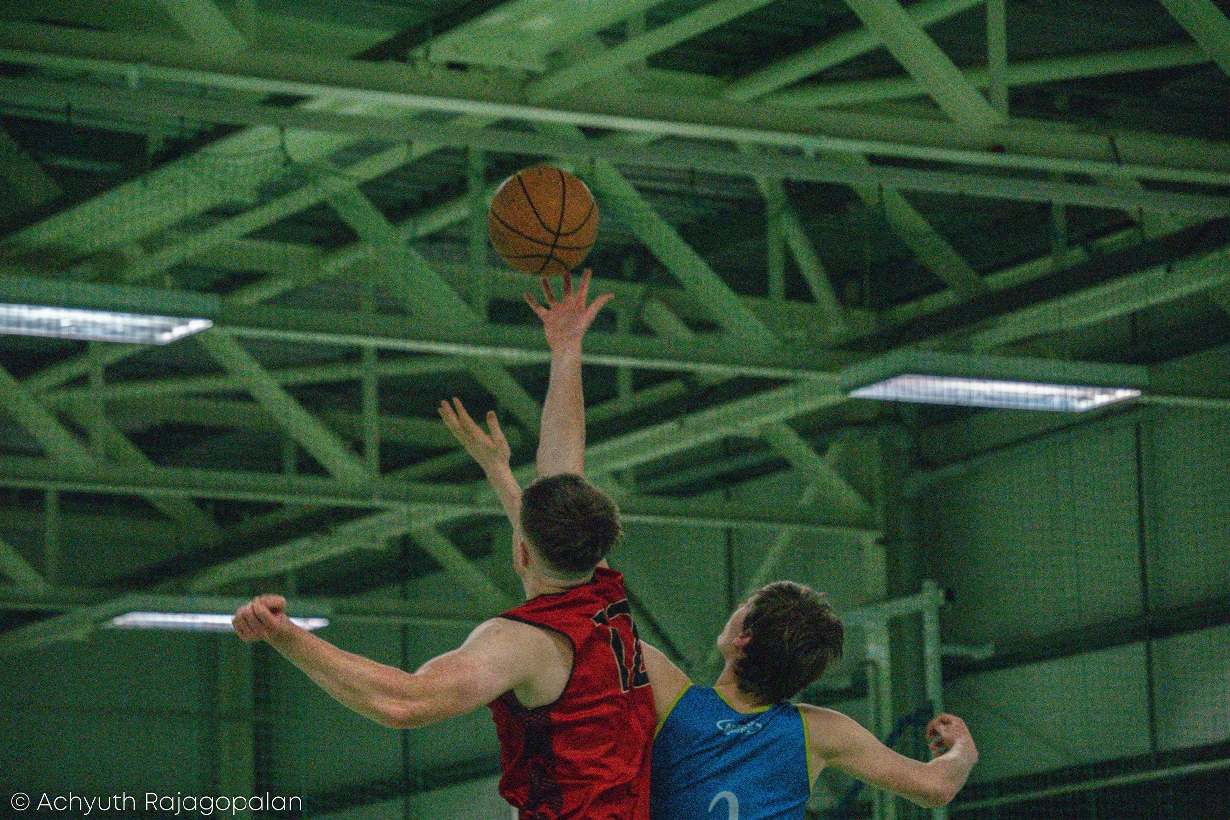 Two young male basketball players, one in a red jersey and the other in a blue jersey, are in action on an indoor basketball court, with the player in red attempting a shot towards the basket while the player in blue tries to defend. The ceiling of t