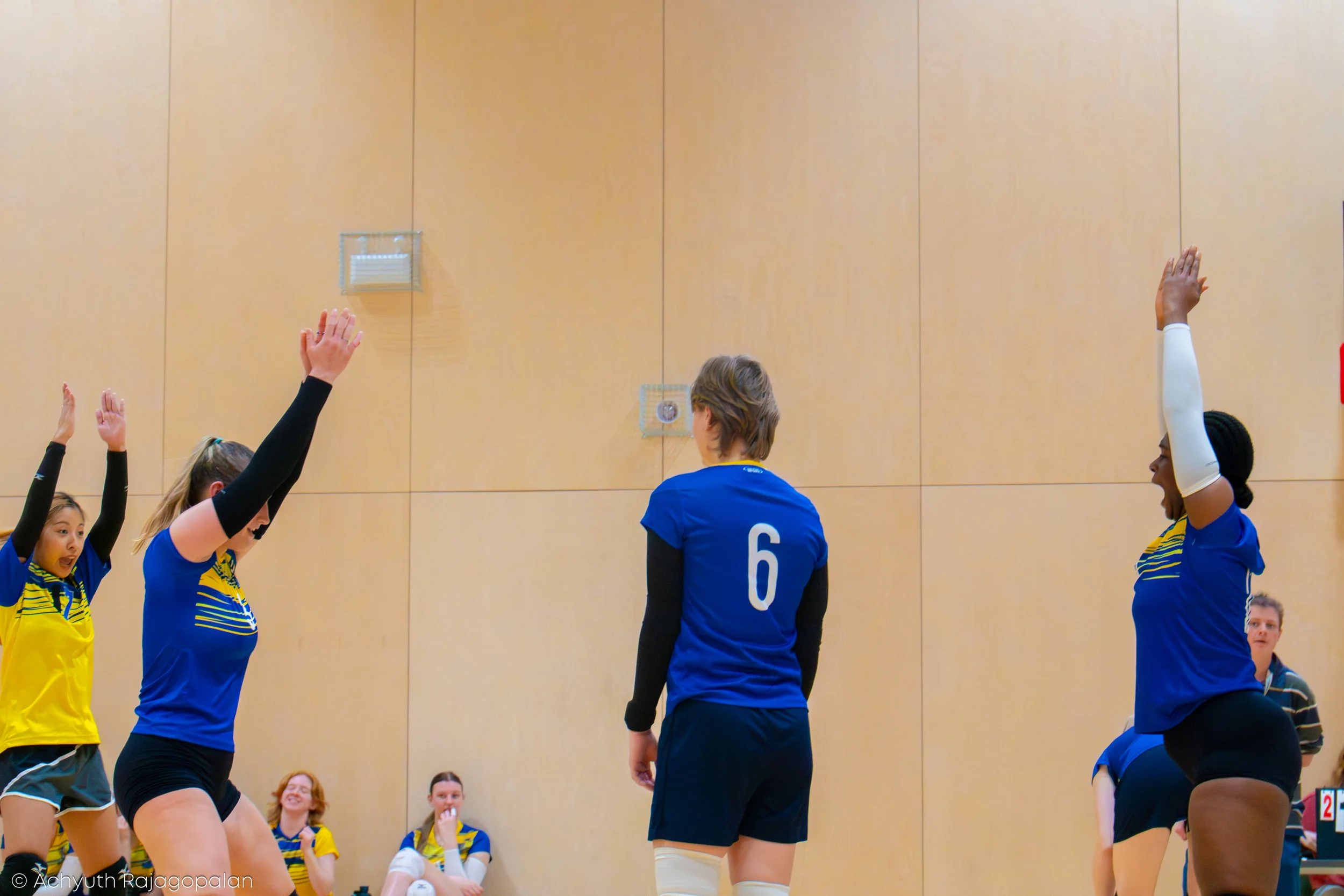 Young female volleyball players in blue and yellow uniforms raising their hands during a game or practice in an indoor gymnasium, with others seated and watching in the background.