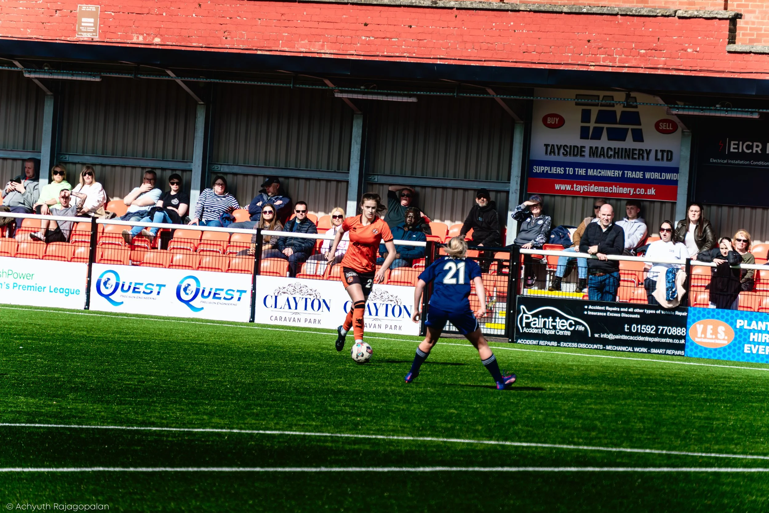 Women and children playing soccer on a green field with spectators sitting on orange and tan seats in the background. A brick building with shops and advertisements is behind the spectators.
