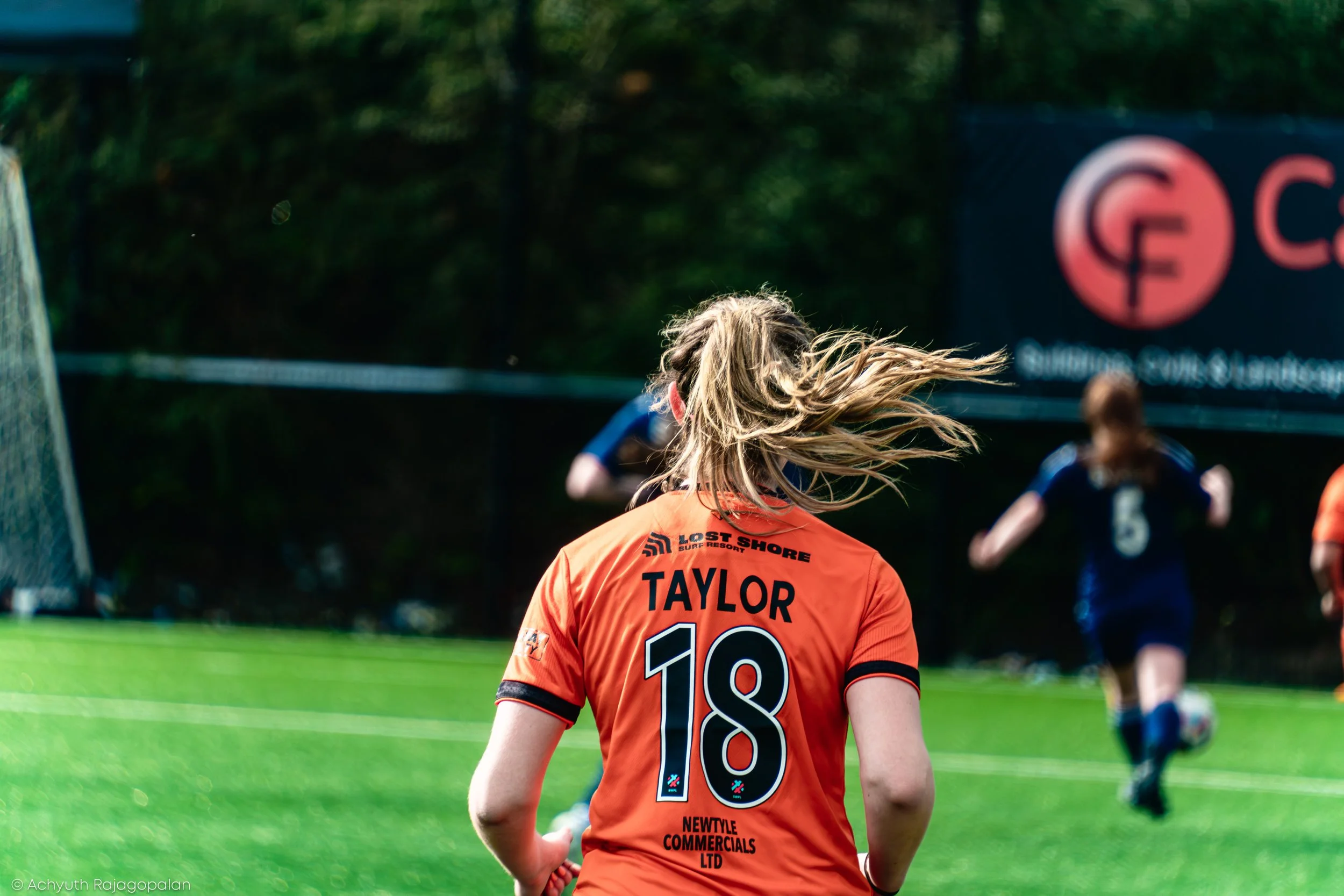 A female soccer player with blonde hair in a ponytail, wearing an orange jersey with the name "TAYLOR" and number 18 on the back, is walking on a soccer field. In the background, other players are visible, and there is a billboard with a logo and ema