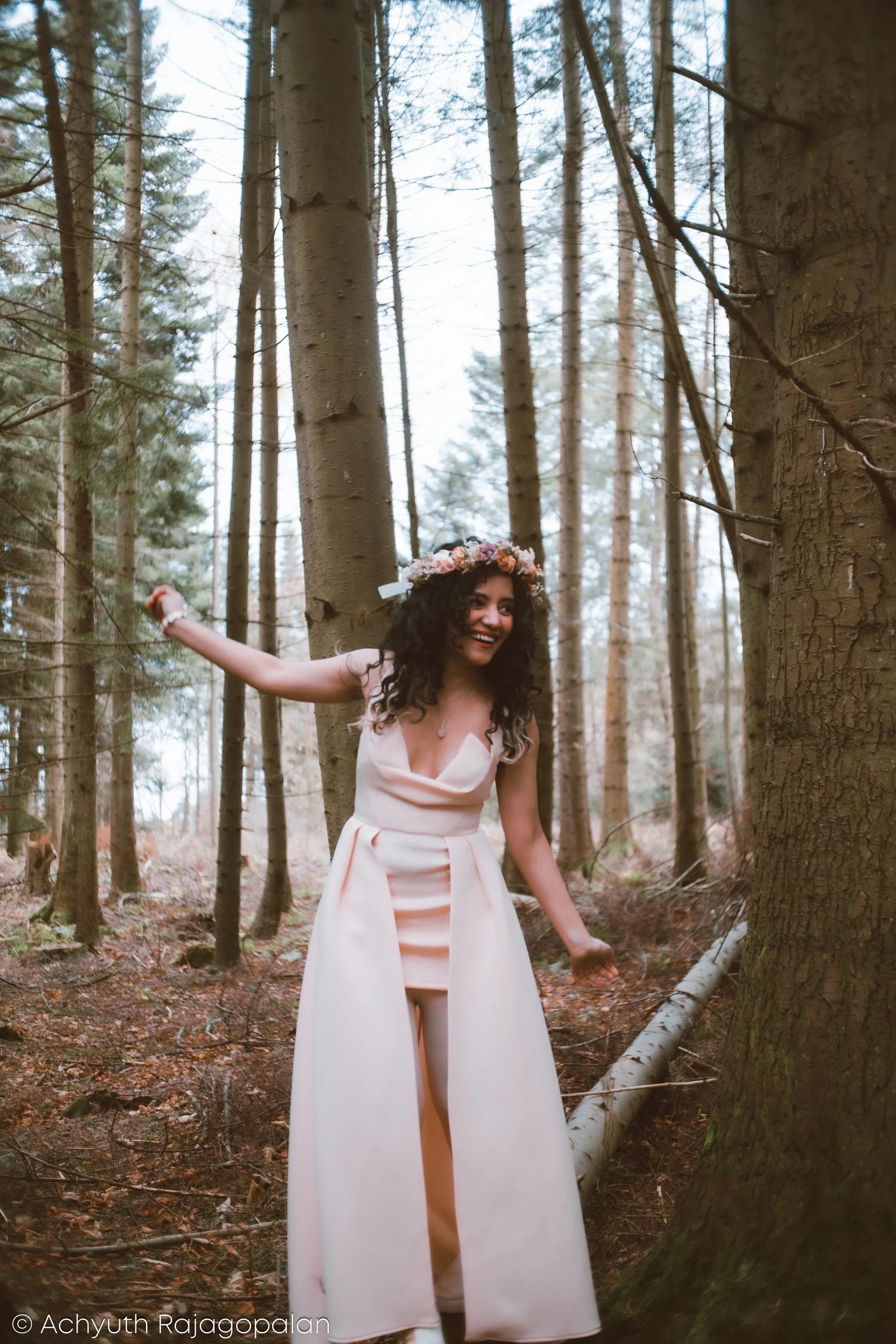 A woman wearing a white dress and a floral crown standing among tall trees in a forest, smiling and extending her arm.