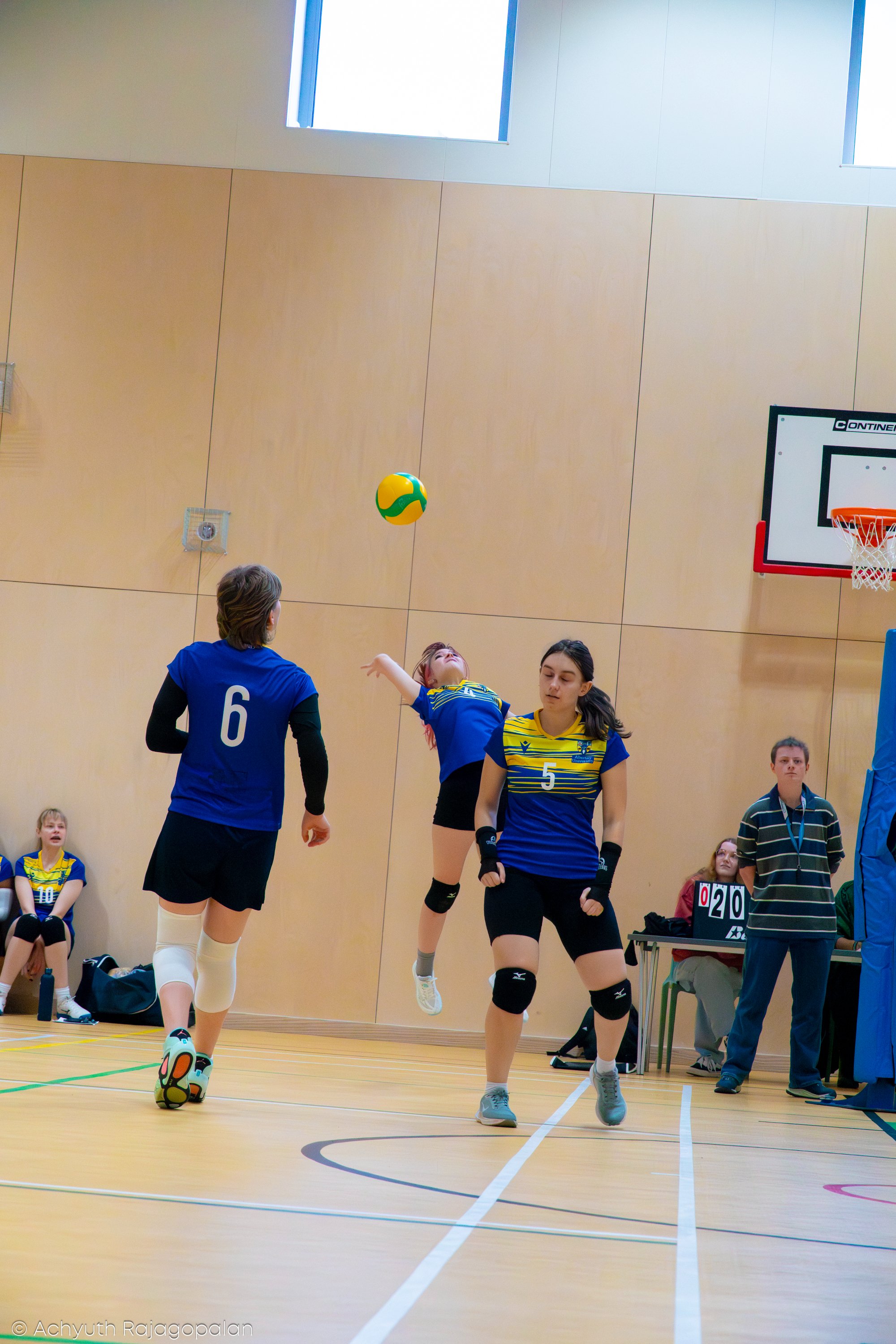 Young girls playing volleyball indoors, with one girl in mid-air preparing to hit the ball, while others observe. Some children are sitting on the floor, and a scoreboard is visible in the background.