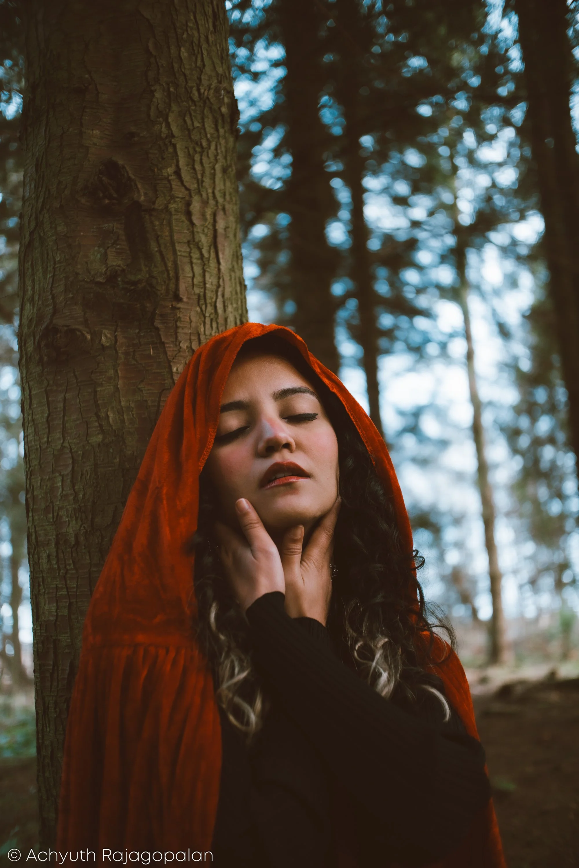 A woman with long curly hair, wearing a red hooded cloak, is standing in a forest with pine trees. Her eyes are closed and her hand is touching her neck.