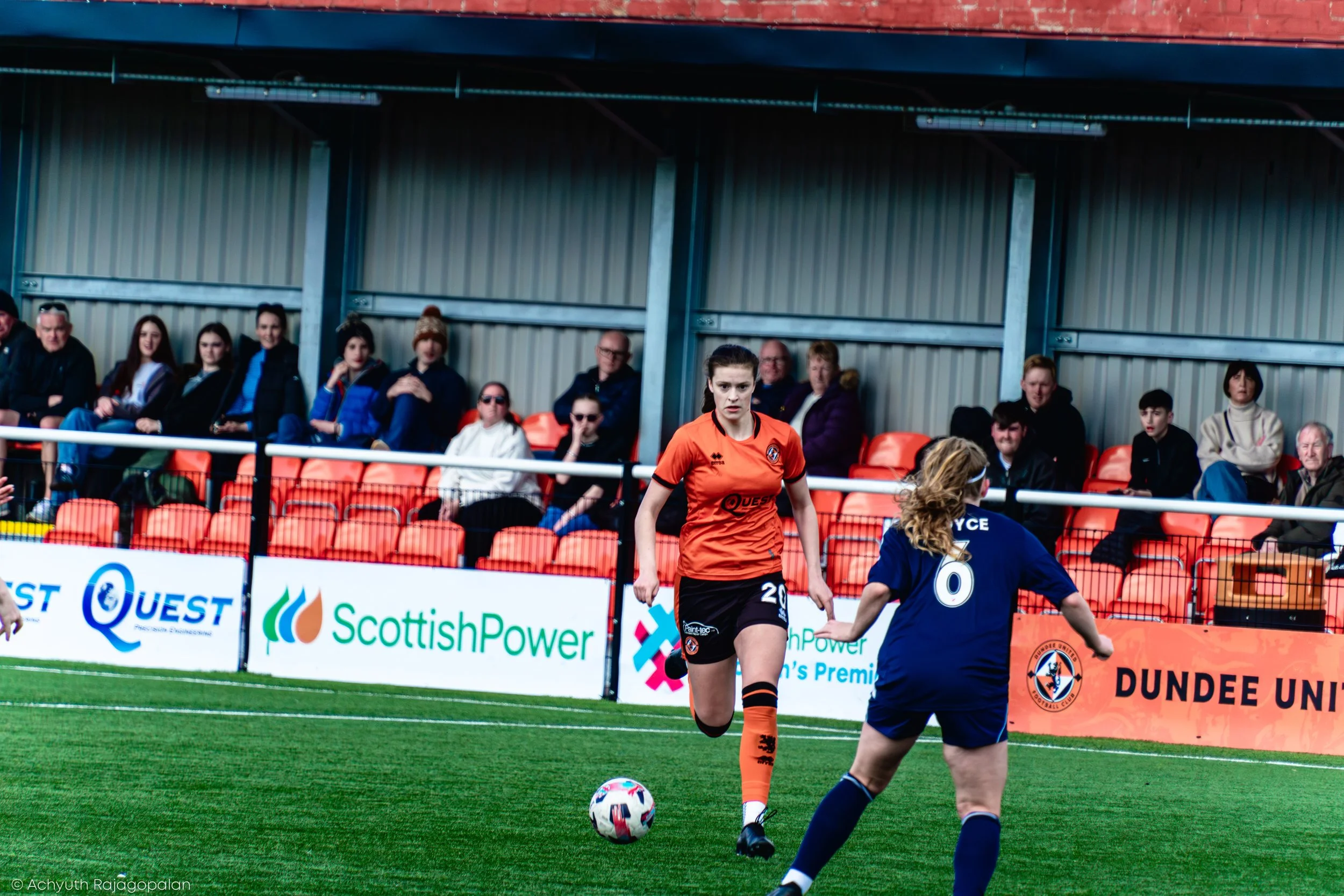 A women's soccer match with a player in a blue jersey controlling the ball on the field, while another player in an orange jersey watches. Spectators sit in the stands behind the field, some wearing winter clothing, with advertising banners along the