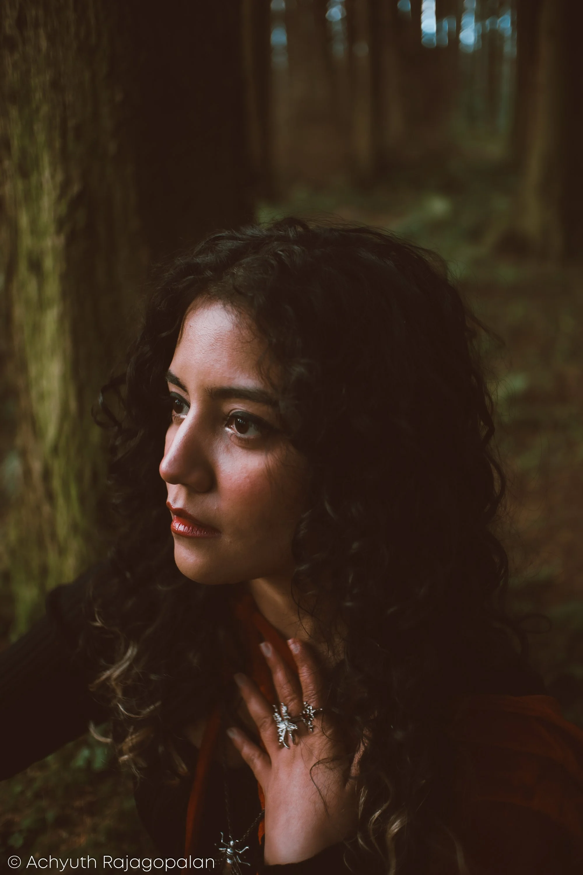 A woman with curly dark hair and light brown skin is outdoors among trees, looking to her left with a serious expression. She is wearing jewelry, including rings and necklaces, and has her hand on her chest.