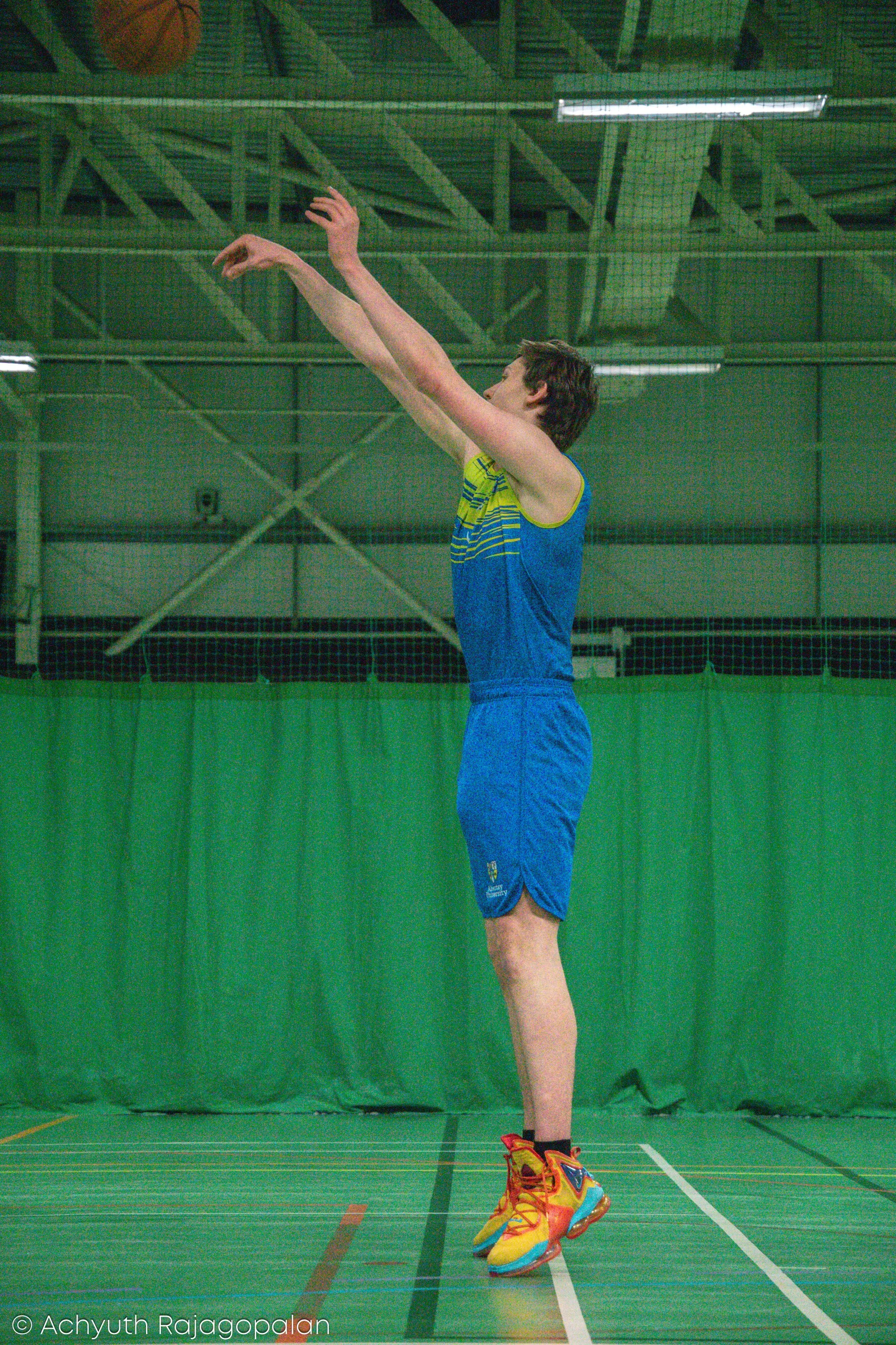 Male athlete in blue sportswear and colorful running shoes standing on a green indoor badminton court, preparing to serve or hit a shuttlecock, with green netting and court boundaries visible in the background.