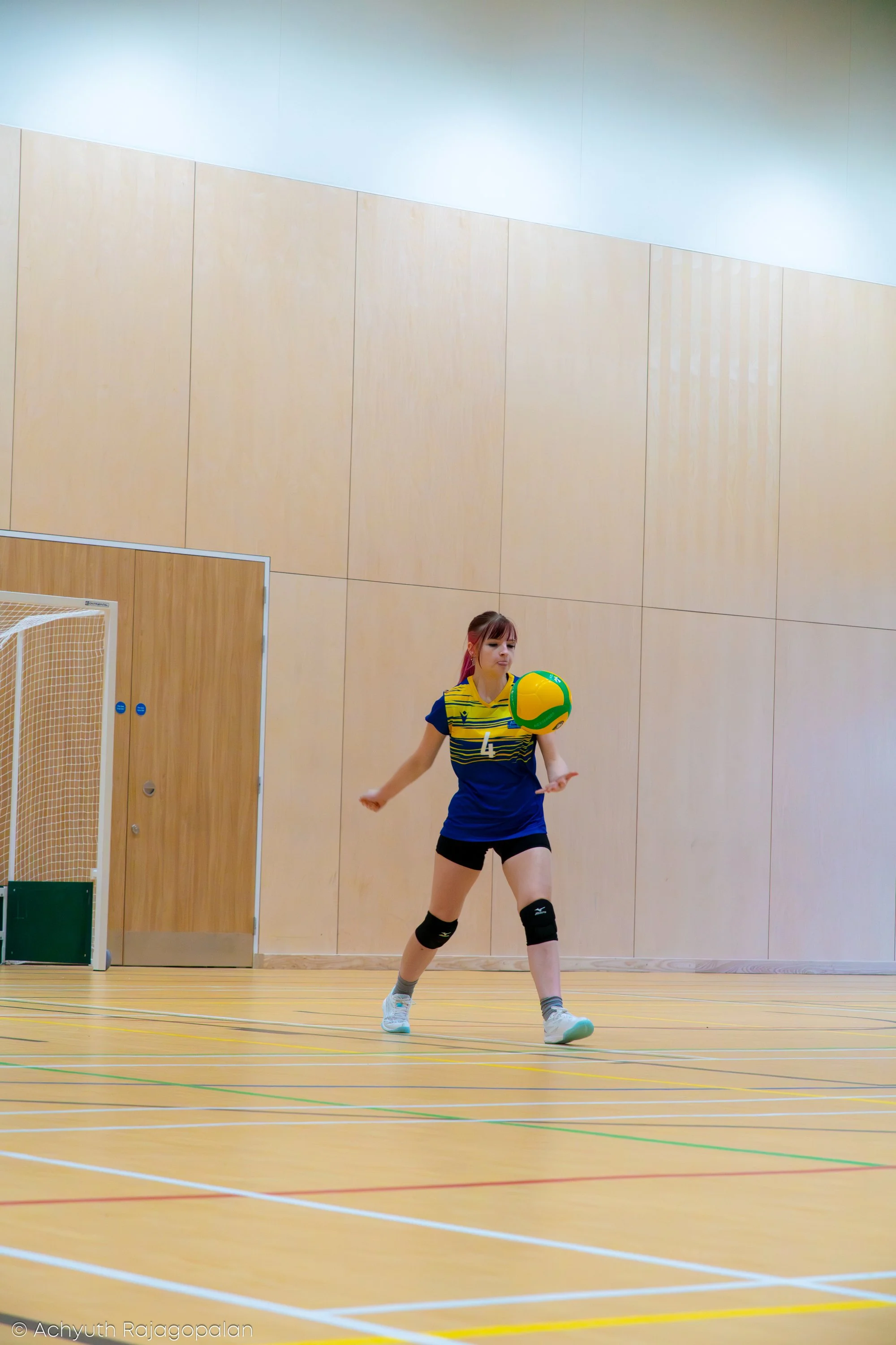 A female volleyball player in a yellow and blue jersey with the number 4, wearing knee pads, playing indoors in a gymnasium with wooden floors and beige paneled walls, appears to be preparing to hit or pass a yellow, green, and white volleyball.