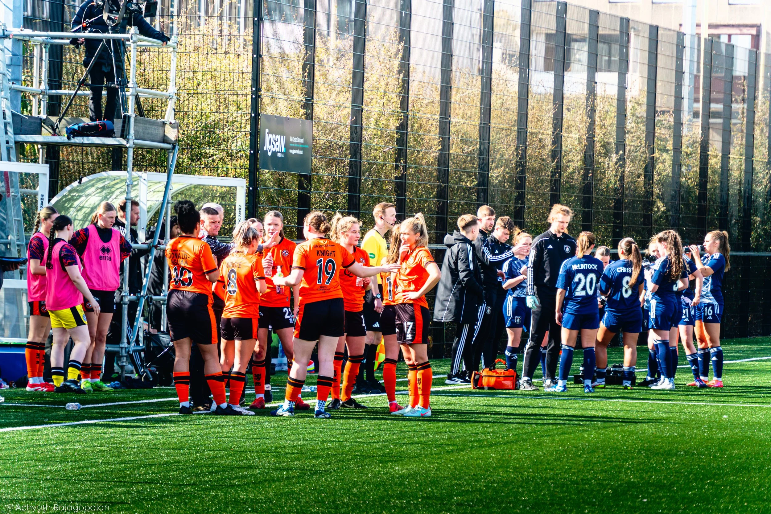 A group of female soccer players standing in a line on the field, wearing various colored uniforms, with a glass building and trees in the background.