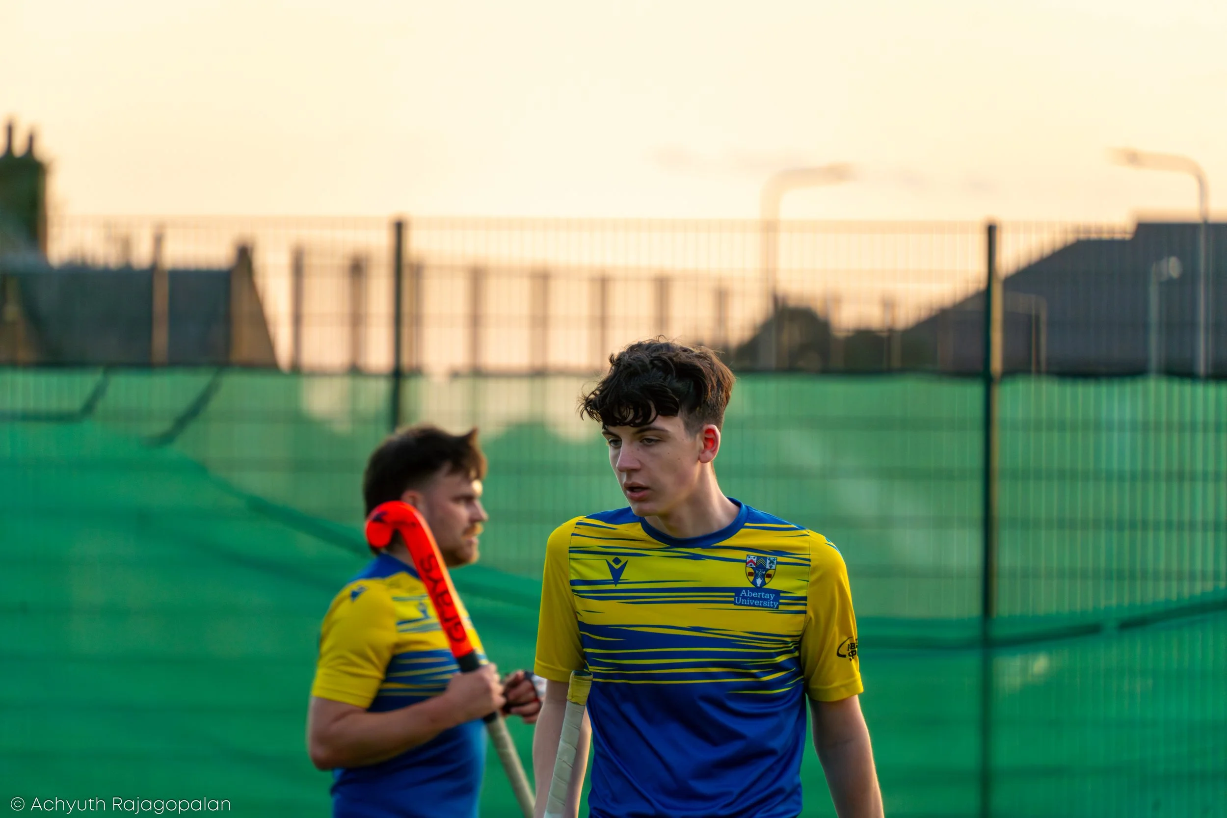 Two young male field hockey players wearing yellow and blue jerseys with the Abertay University logo, standing in front of a green fence at a sports field during sunset, with industrial chimneys in the background.