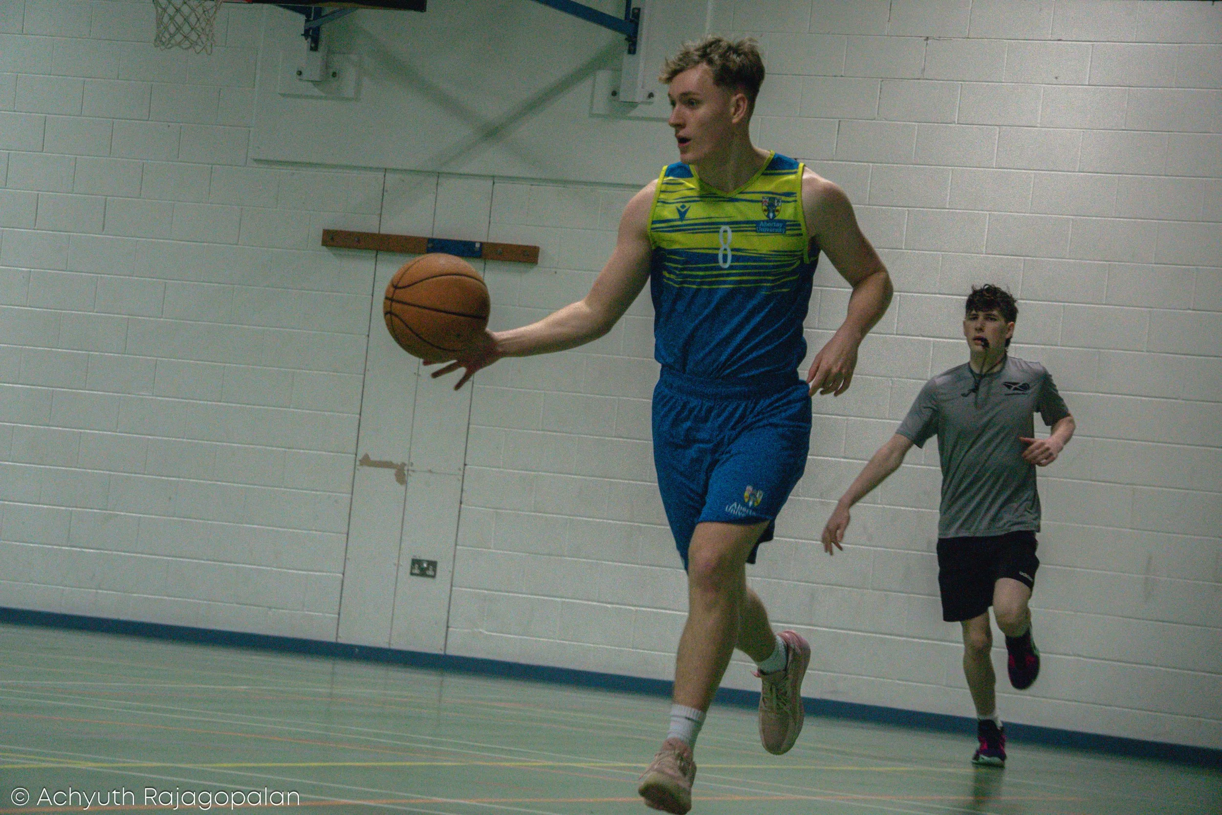 A young male athlete in a multicolored sports uniform dribbles a basketball during an indoor game, with another young player running behind him.