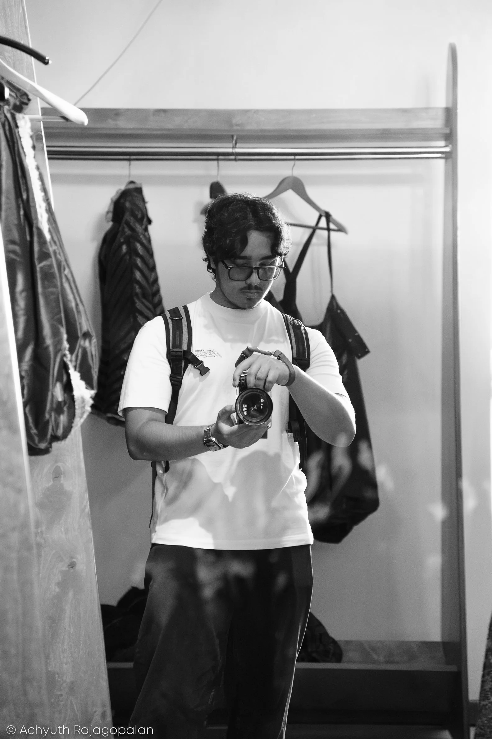 A young man with curly hair, glasses, and a mustache taking a selfie in a mirror while wearing a t-shirt and backpack. Behind him, a clothing rack with hangers and a few garments is visible, and the setting appears to be a room or closet.