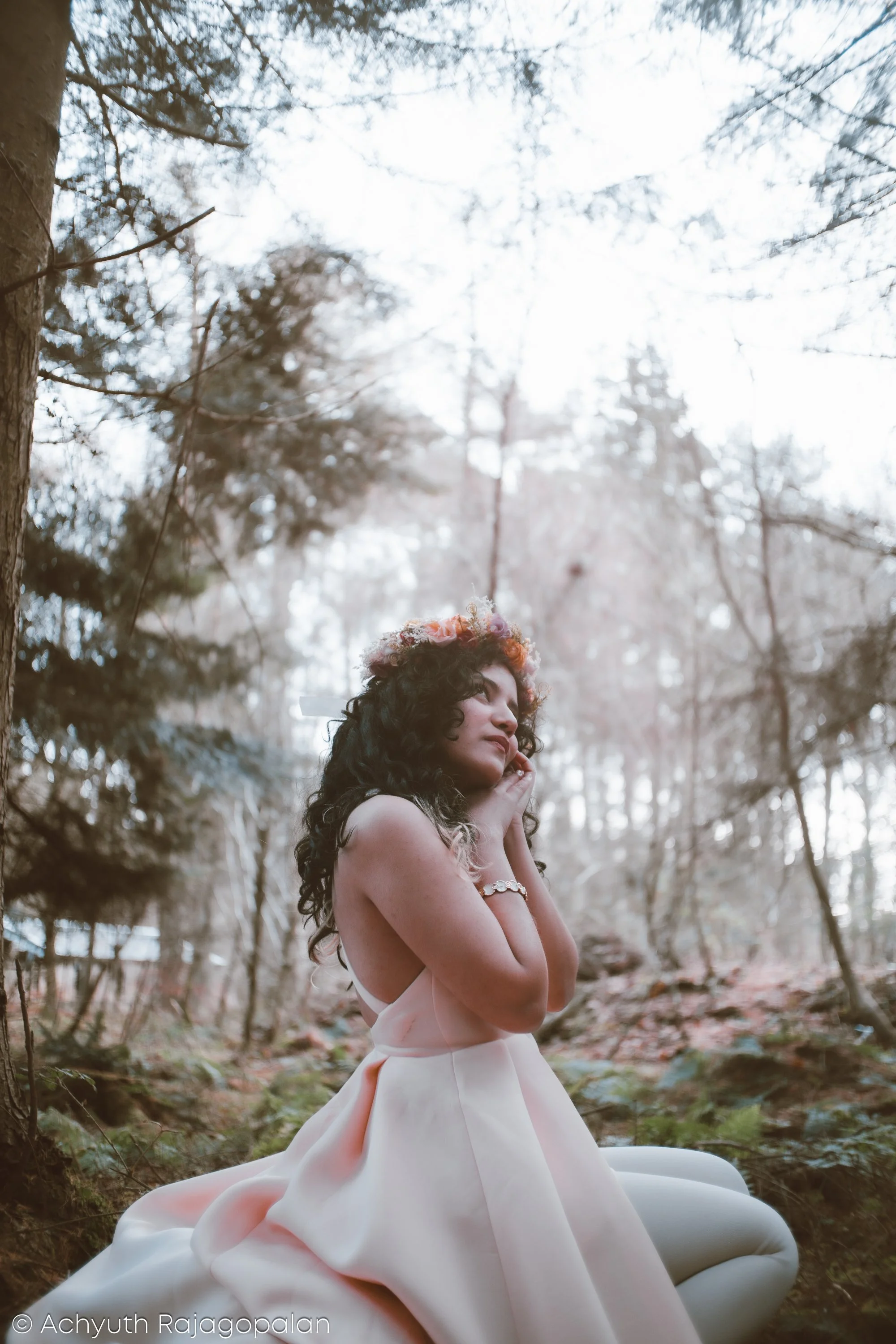 A woman with curly hair wearing a flower crown and a strapless dress, standing outdoors in a forest with tall trees and sunlight filtering through the branches.