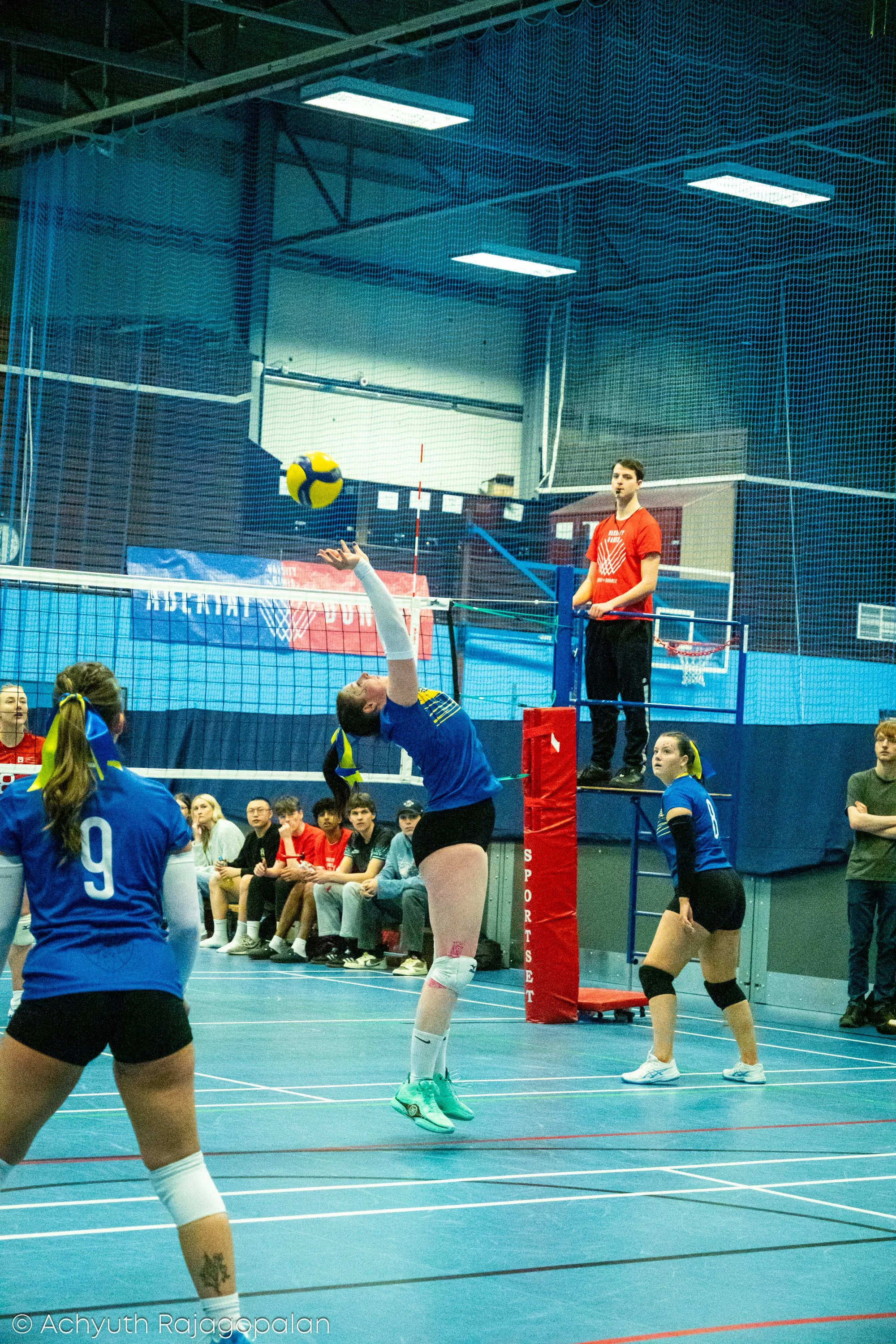 A volleyball game in progress with players on a court, one player jumping to block a ball, others observing and preparing to react, spectators sitting on the sidelines, gymnasium with blue floor and net, scoreboard, and referee standing on a raised p