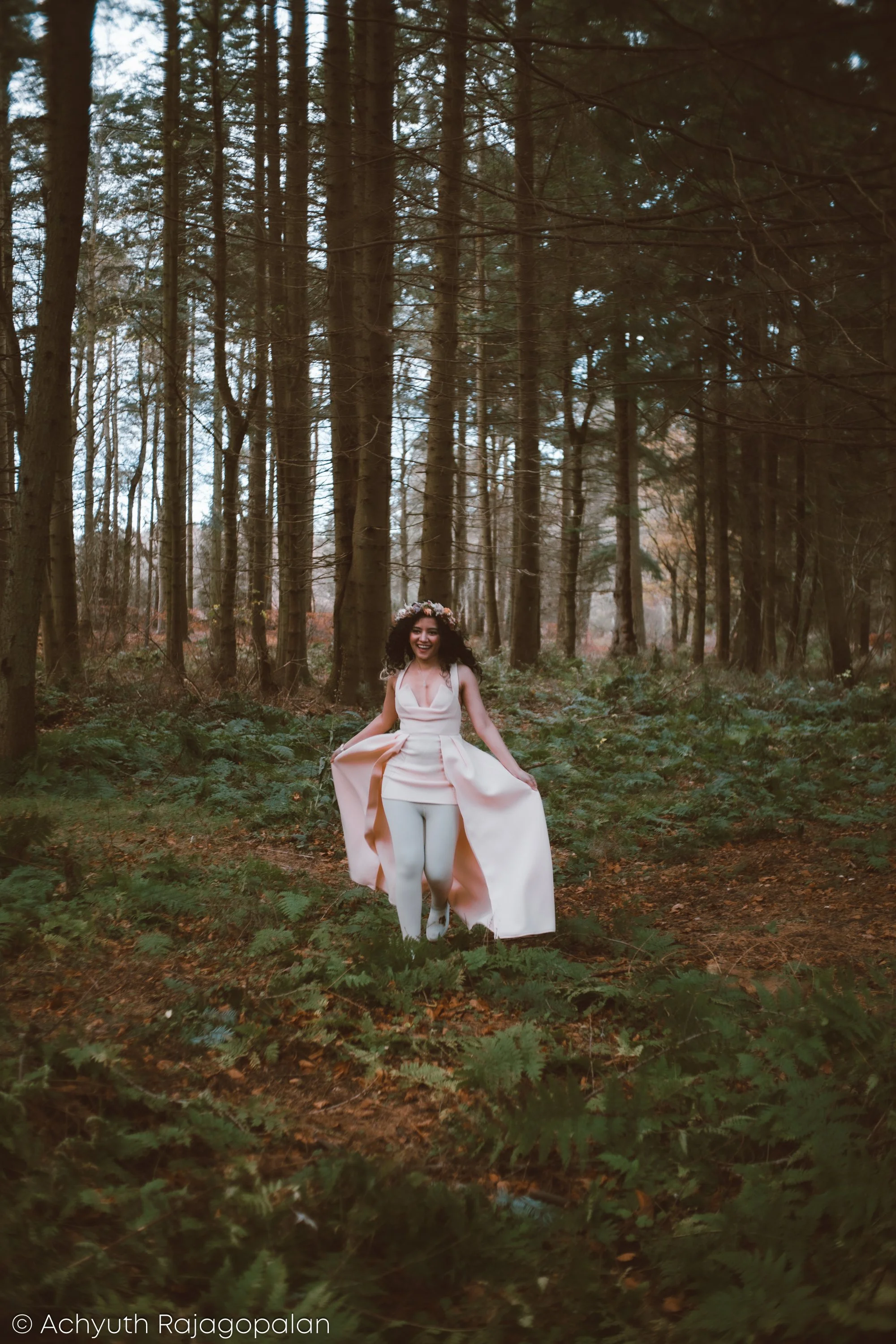 A woman wearing a light pink dress and white leggings is sitting on the ground in a forest surrounded by tall trees and green foliage.