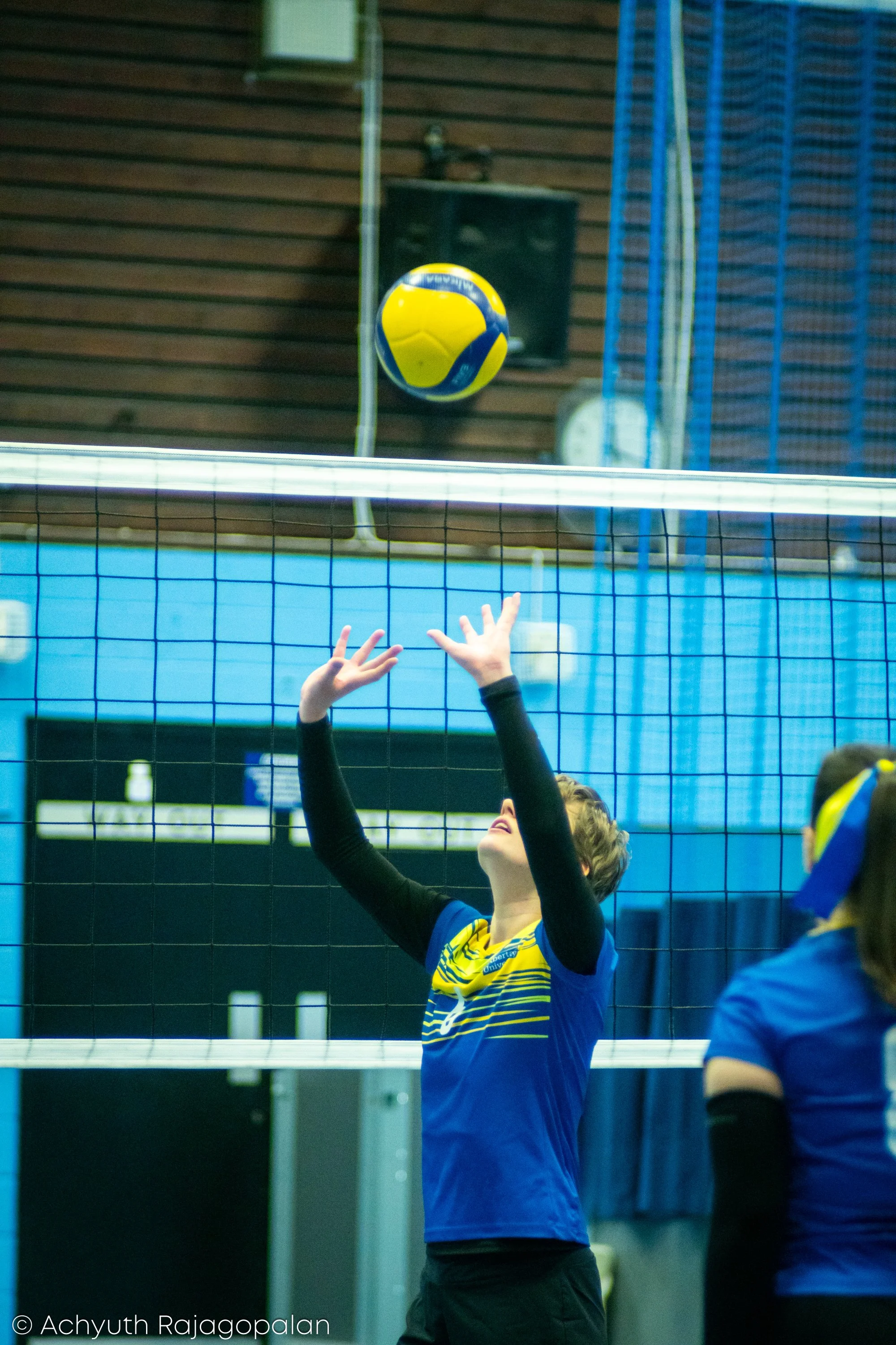 A volleyball player in blue and yellow with arms raised, preparing to hit a volleyball in an indoor gym.