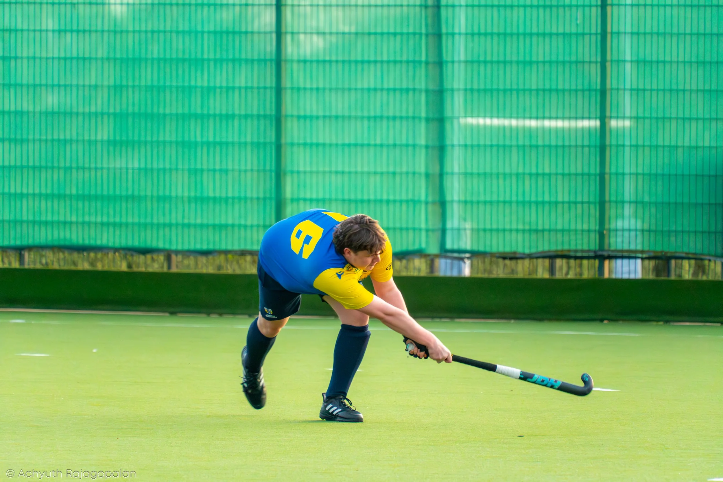 A young male field hockey player in a blue and yellow uniform is bent over and reaching down to pick up a field hockey ball with his stick on a green field, with a green fence in the background.
