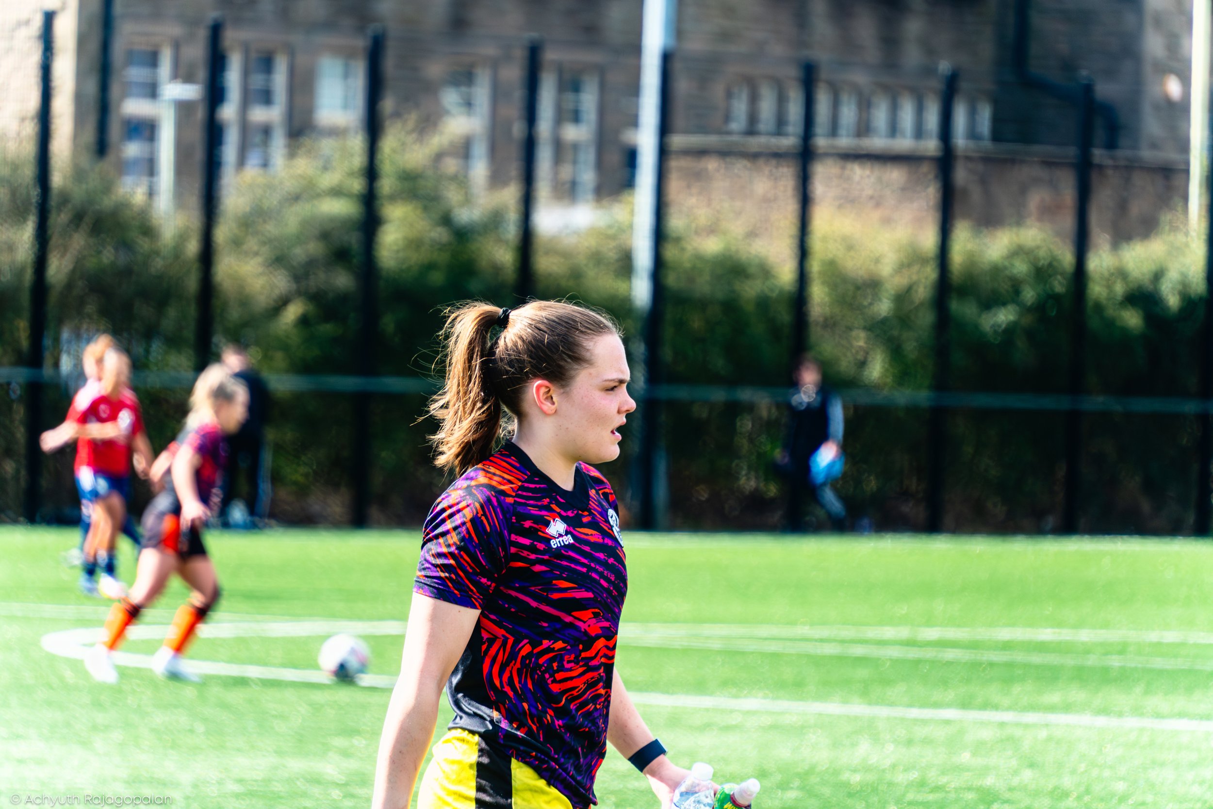 A female soccer player with a ponytail wearing a colorful jersey and yellow shorts, standing on the field during a game with other players in the background.