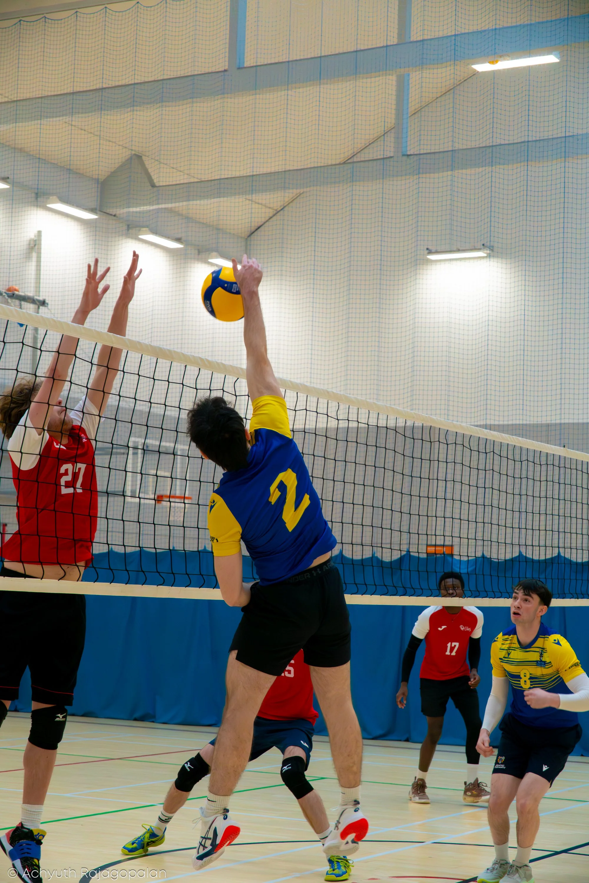 Indoor volleyball game with players jumping and reaching for the ball near the net, some players wearing red uniforms and others in blue and yellow uniforms, in a gymnasium with bright lighting and a high ceiling.