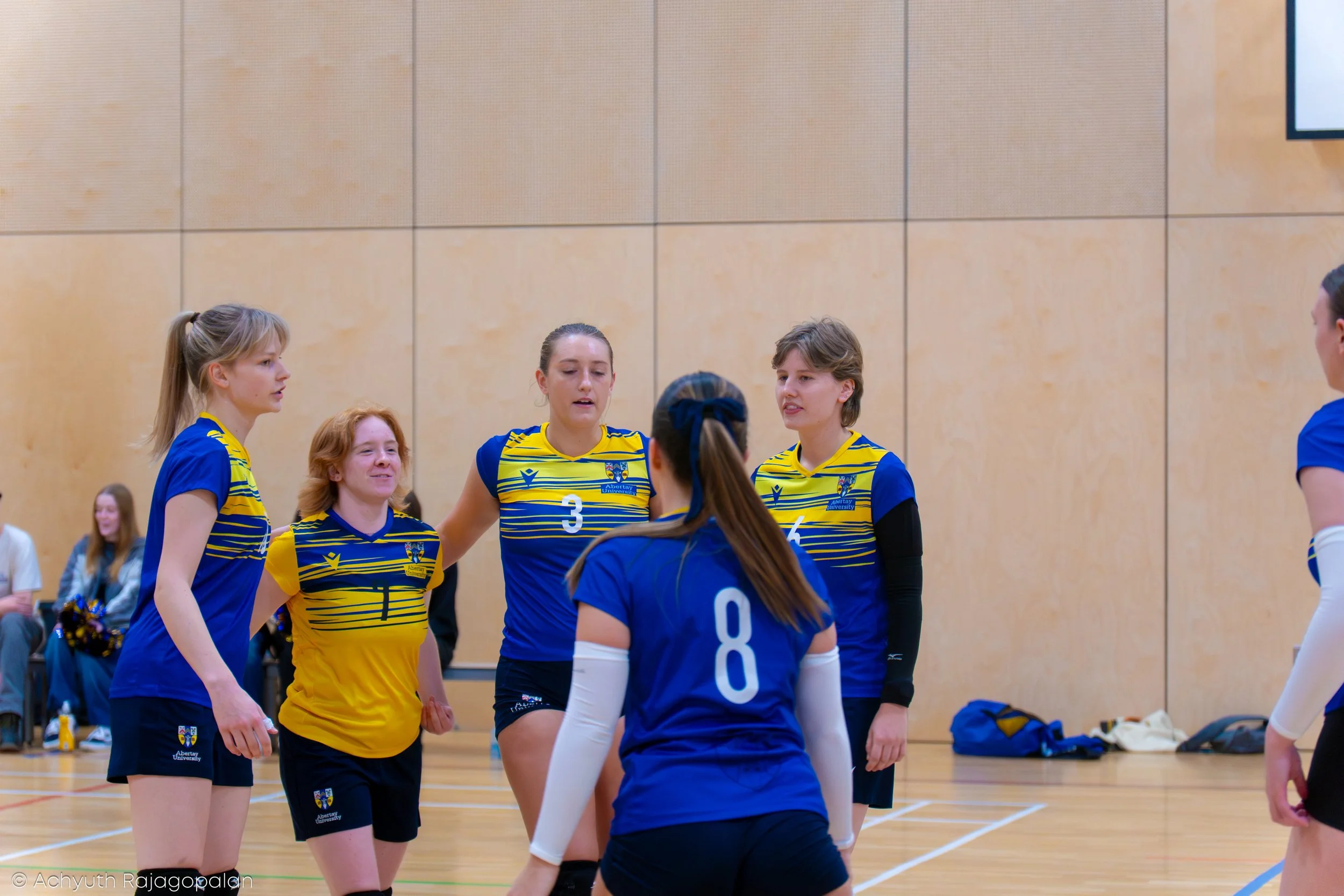 A group of female volleyball players in blue and yellow uniforms gathered on an indoor court, likely during a match or team meeting.
