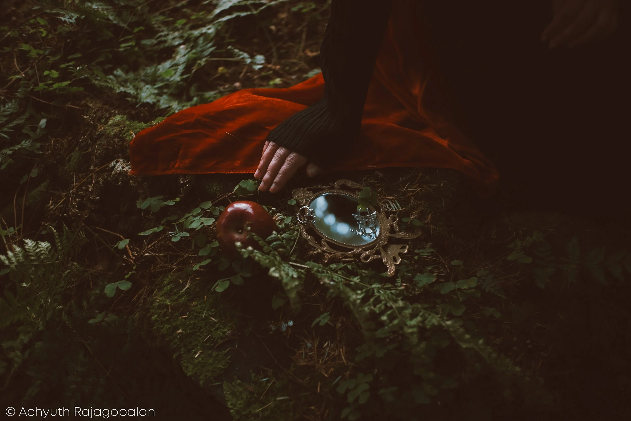 Person placing an ornate hand mirror on the ground in a forest, near an apple, with a reddish cloth underneath.