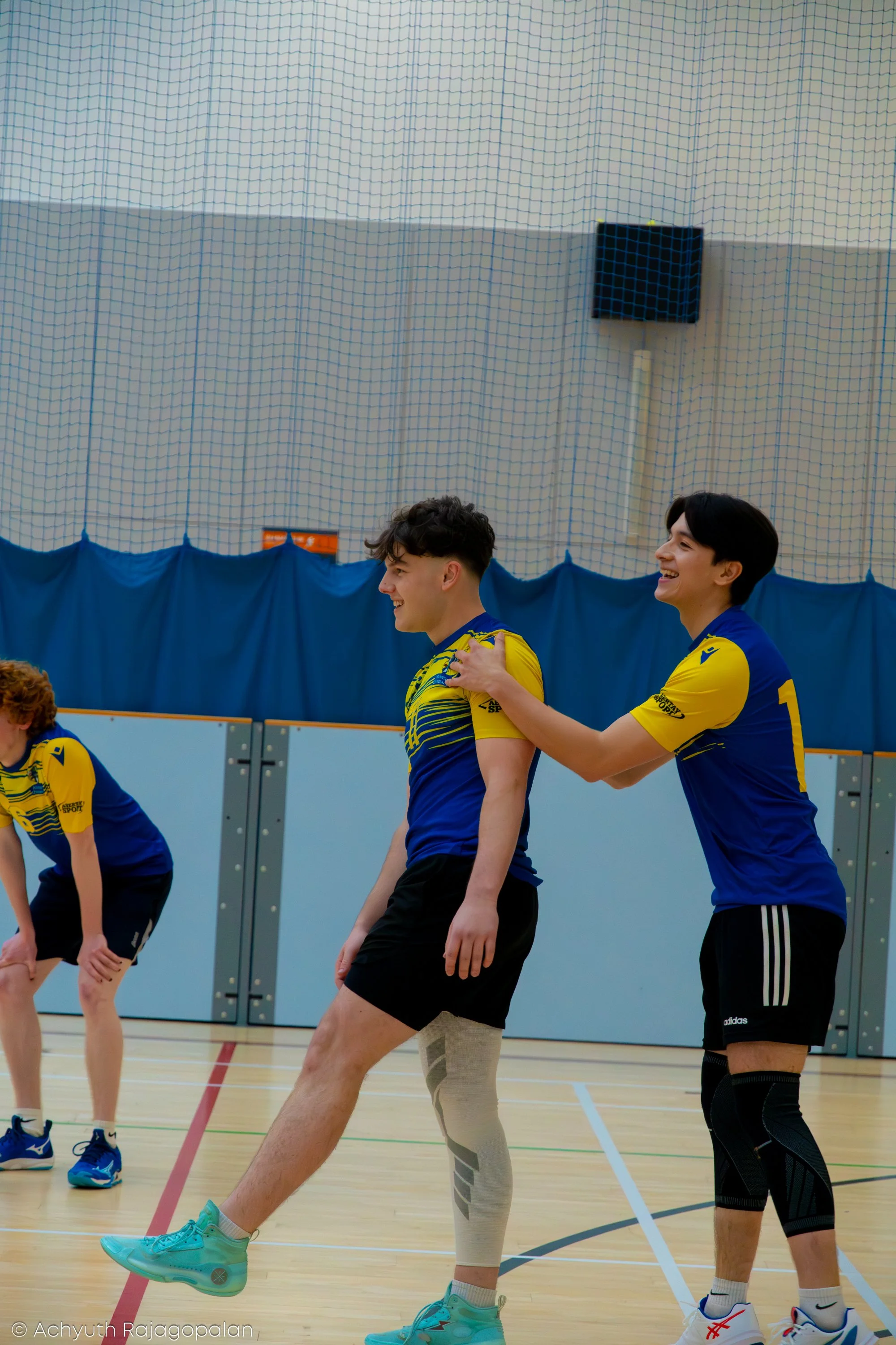 Two young men in athletic uniforms stretching in a gymnasium, with a net and a speaker in the background.