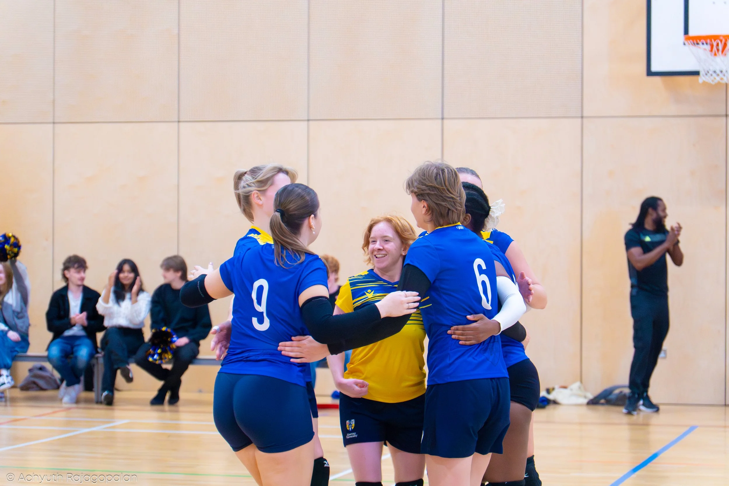 Group of female volleyball players embracing each other after a game in a gymnasium, with spectators seated on benches in the background.