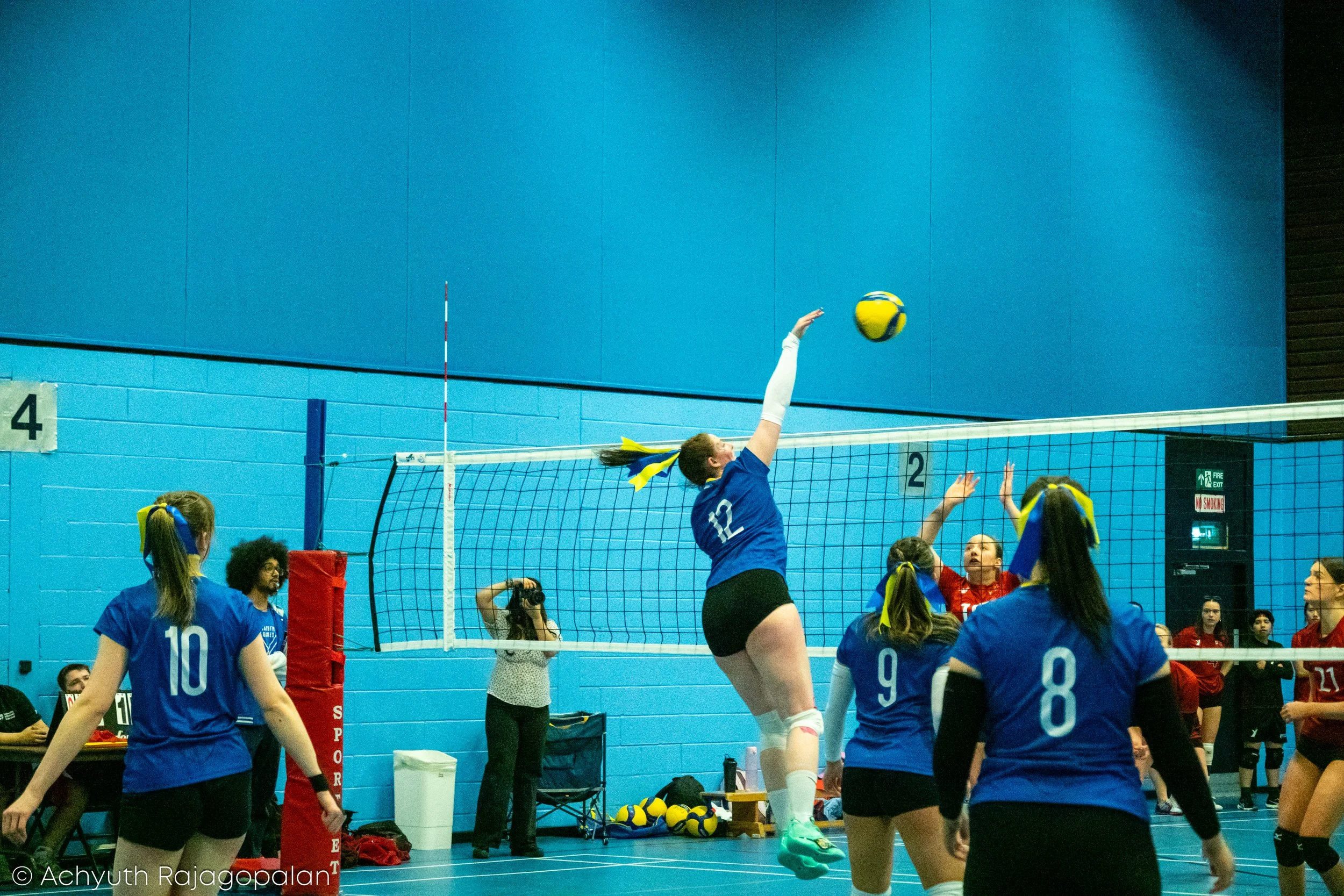 Female volleyball players in blue jerseys jump to hit the ball over the net as teammates and opponents watch on during a match in an indoor gym.