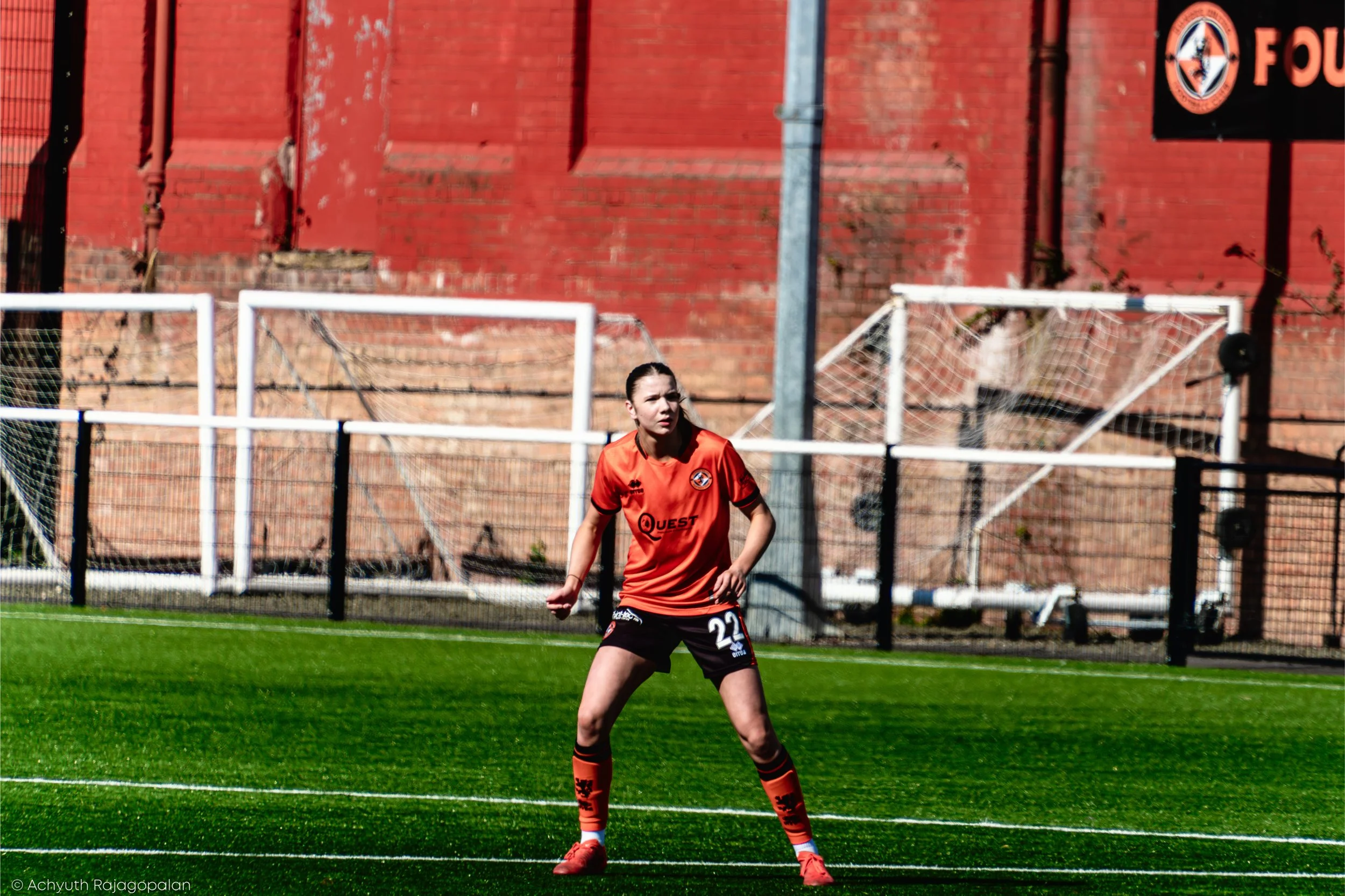 A female soccer player in an orange and black uniform stands on a soccer field with a focused expression, against a background of a red brick wall and soccer goalposts.