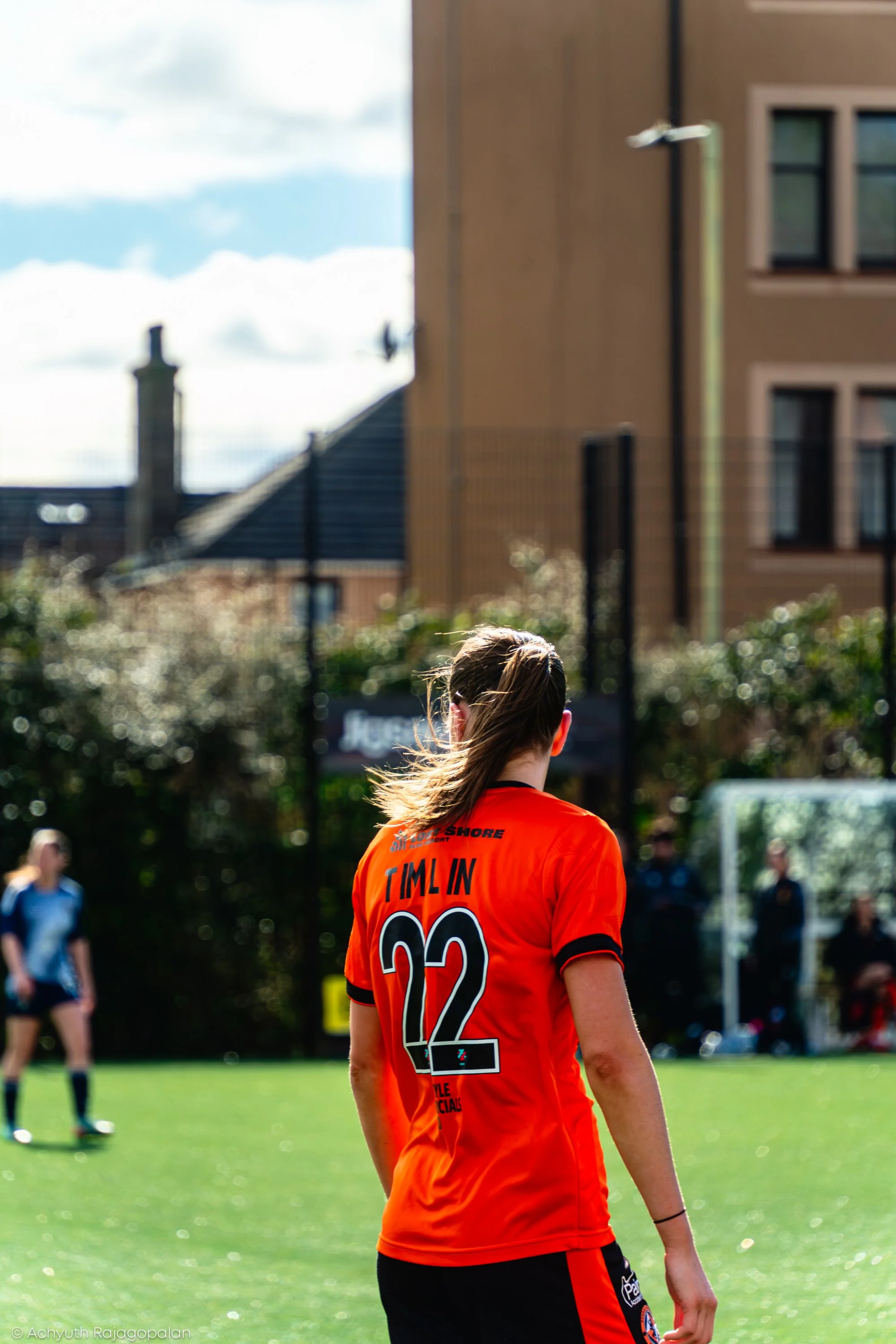 A person in an orange sports jersey with the name 'Tim Lin' and the number 22 on the back, standing outdoors on a grassy field during a sunny day, with other people in the background and buildings nearby.
